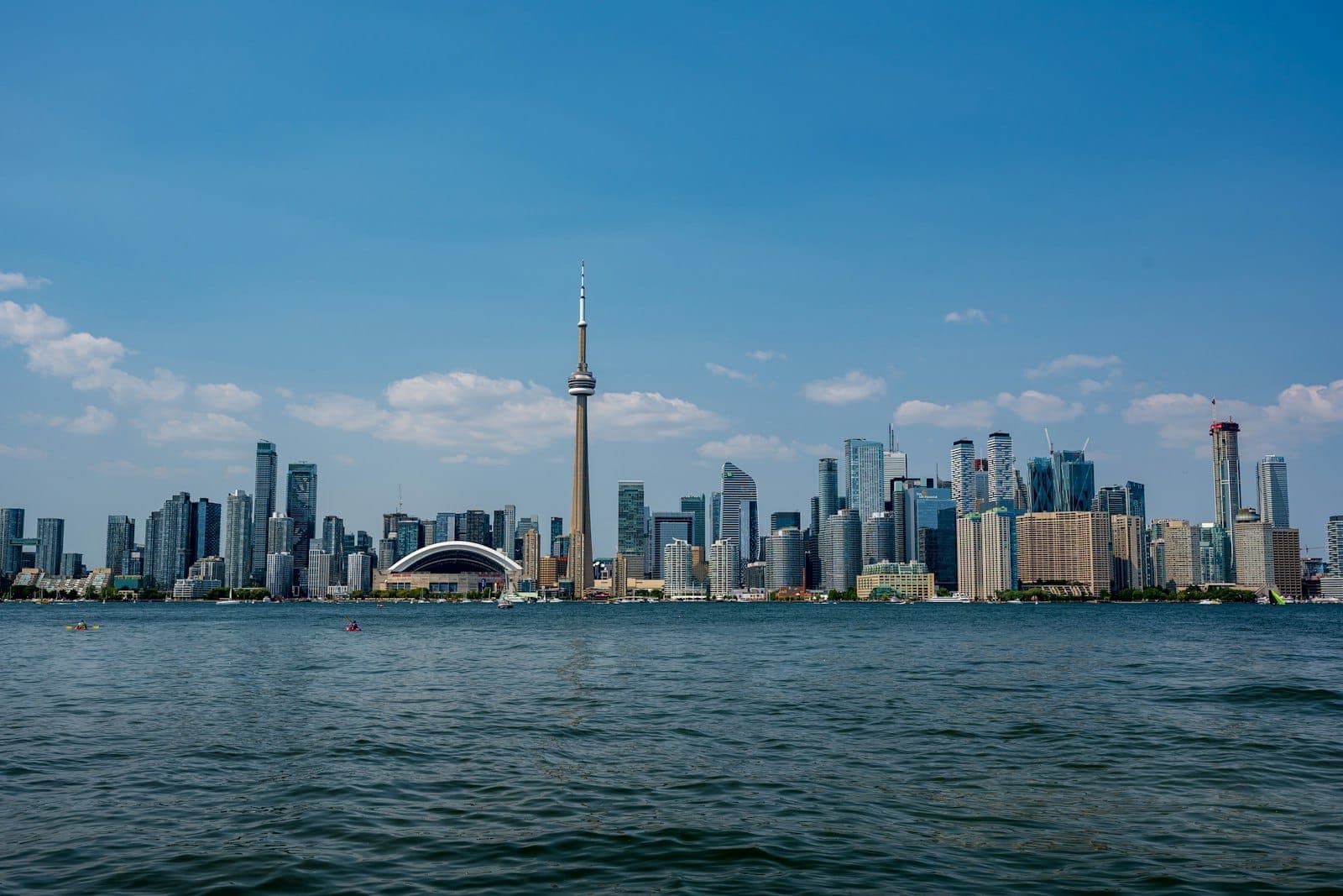 Skyline di Toronto con la CN Tower in una giornata di sole, vista dall'acqua con cielo azzurro e poche nuvole.