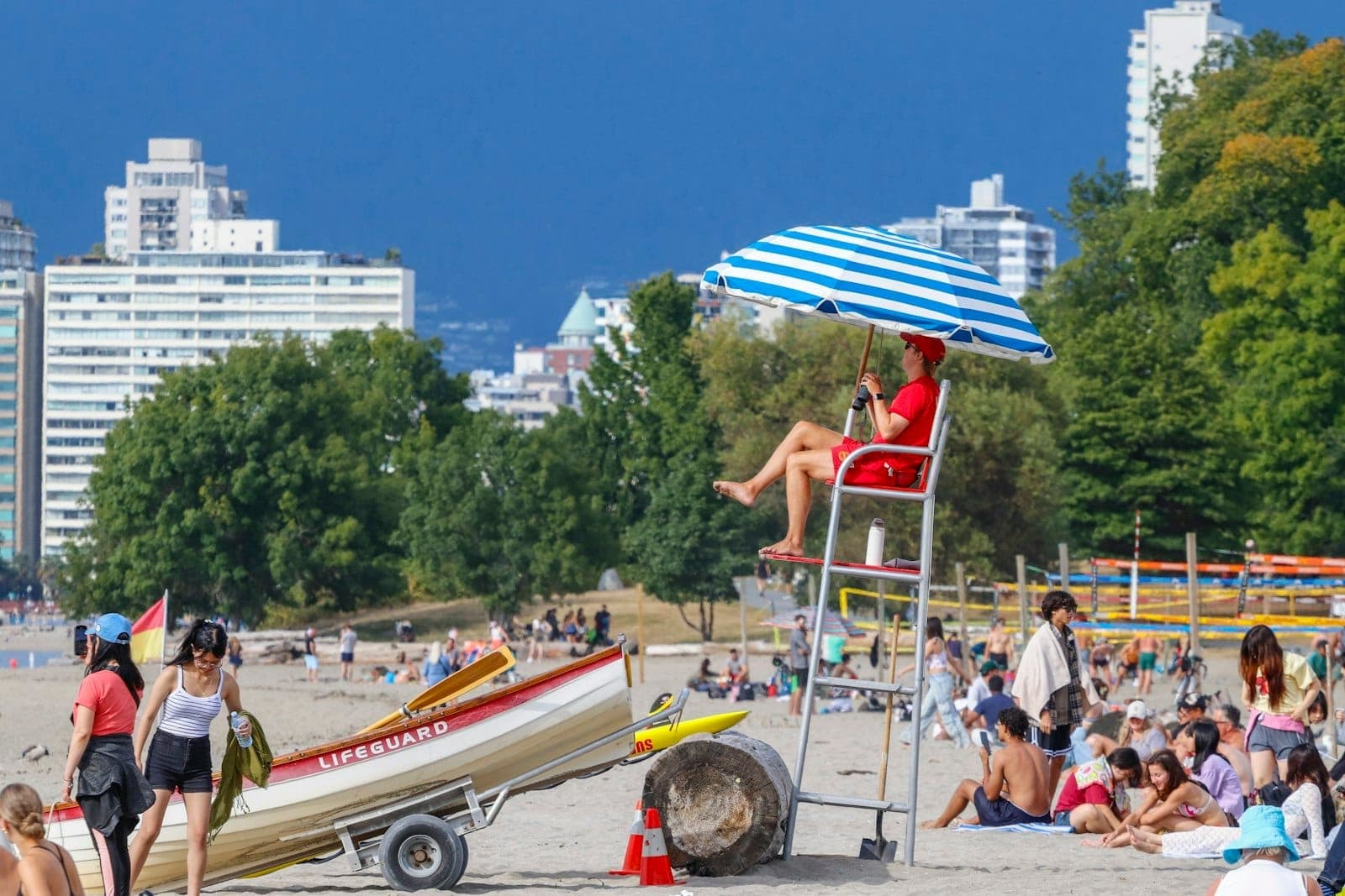 ชายหาด Vancouver ที่คึกคักพร้อมเจ้าหน้าที่กู้ภัย นักอาบแดด และอาคารในเมืองในช่วงฤดูร้อนที่มีอากาศแจ่มใส