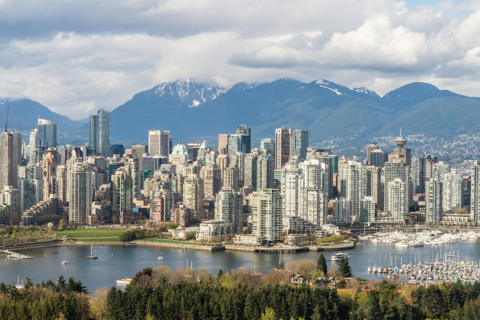 ขอบฟ้าเมือง Vancouver พร้อมอาคารสมัยใหม่ ทางน้ำ มารีนา ต้นไม้เขียวชอุ่ม และภูเขายอดหิมะภายใต้ท้องฟ้าที่มีทั้งเมฆและแสงแดด