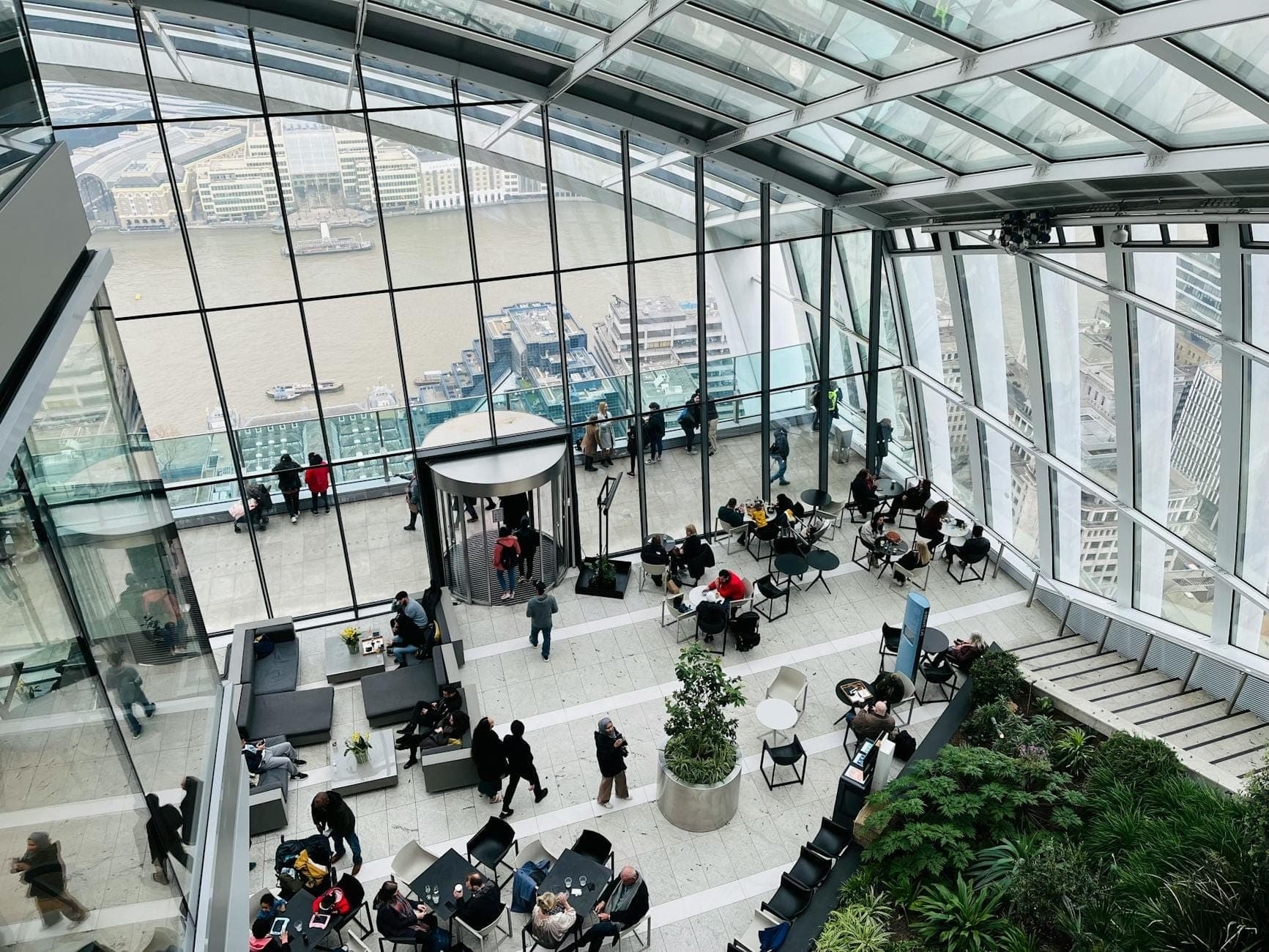 Vista interna di un grande giardino pensile in vetro a Londra con rigogliosa vegetazione, persone che godono delle viste sulla città e il Tamigi attraverso finestre panoramiche.