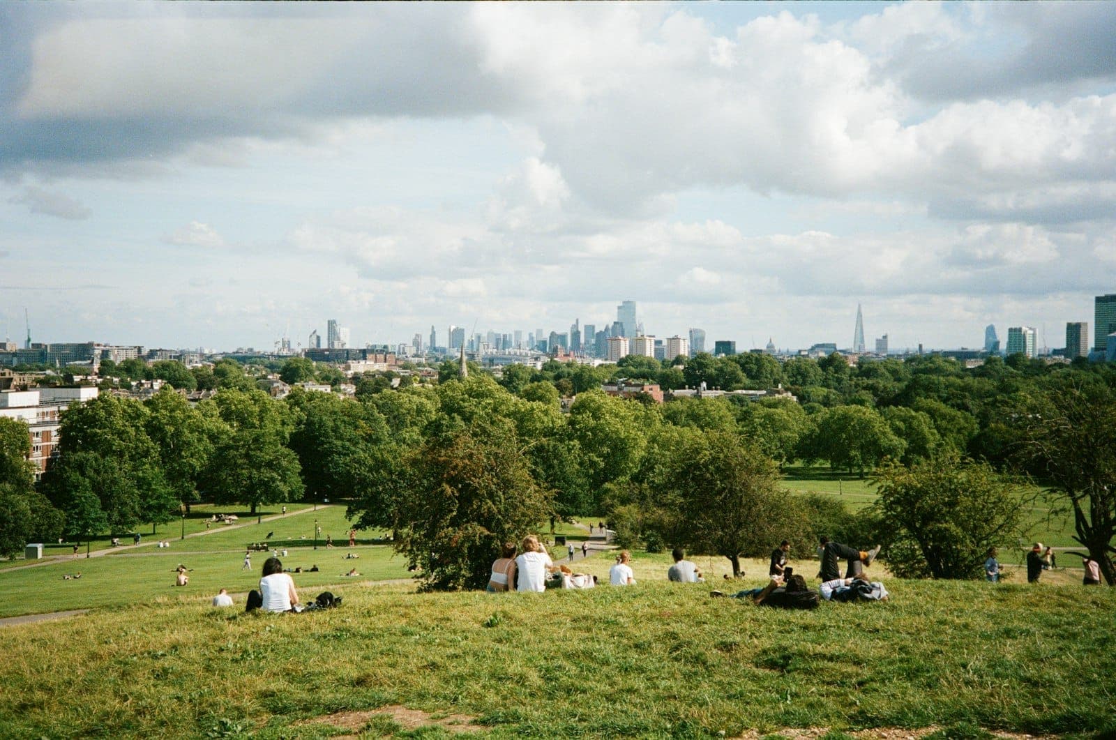 Vista panoramica dalla cima di una collina erbosa in un parco di Londra, con persone che si rilassano e lo skyline della città e il verde sullo sfondo.