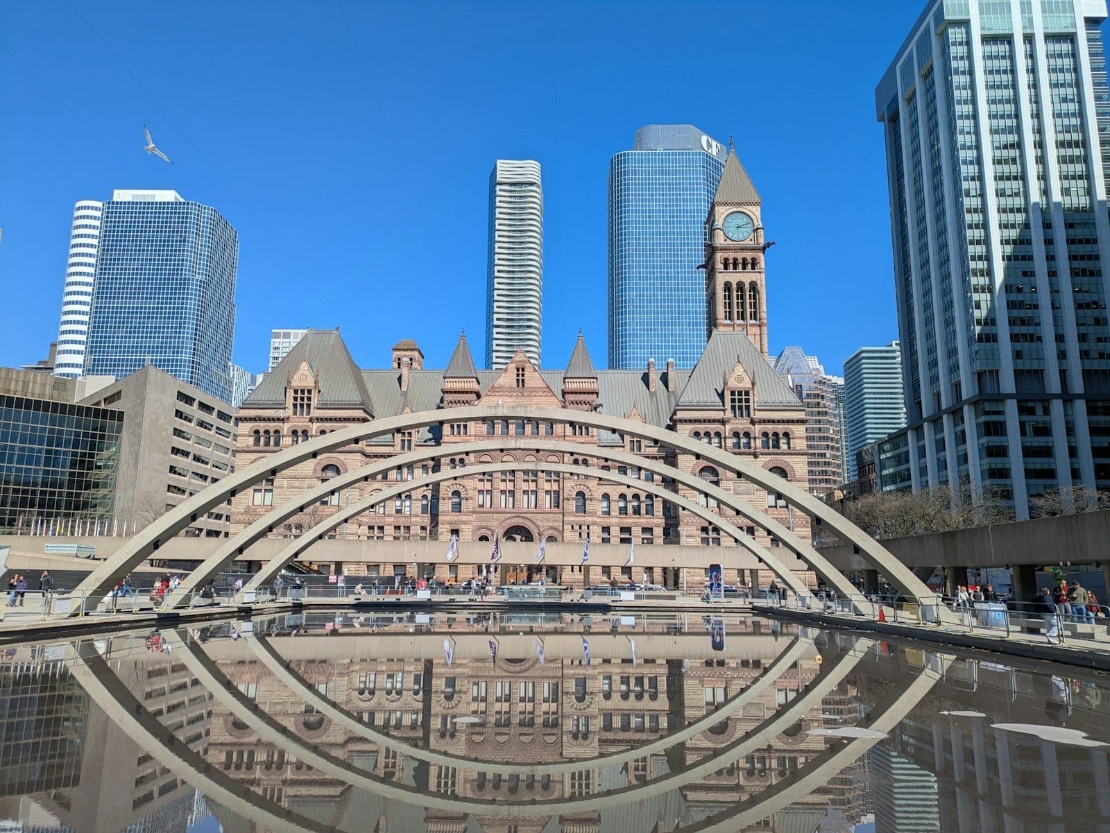 La Nathan Phillips Square de Toronto con sus icónicas arcadas, estanque reflectante, torre del reloj del Old City Hall y modernos rascacielos bajo un cielo azul despejado.