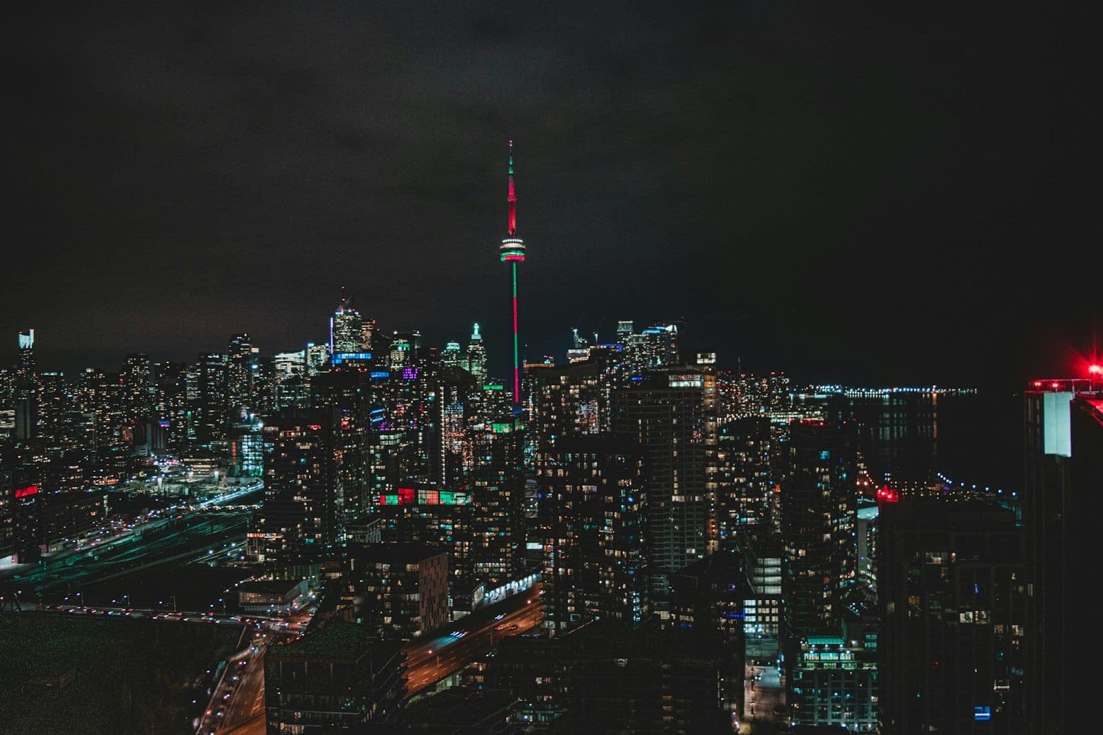 Cakrawala Toronto di malam hari dengan CN Tower yang bercahaya di tengah, dikelilingi gedung-gedung pencakar langit yang menyala di bawah langit gelap.