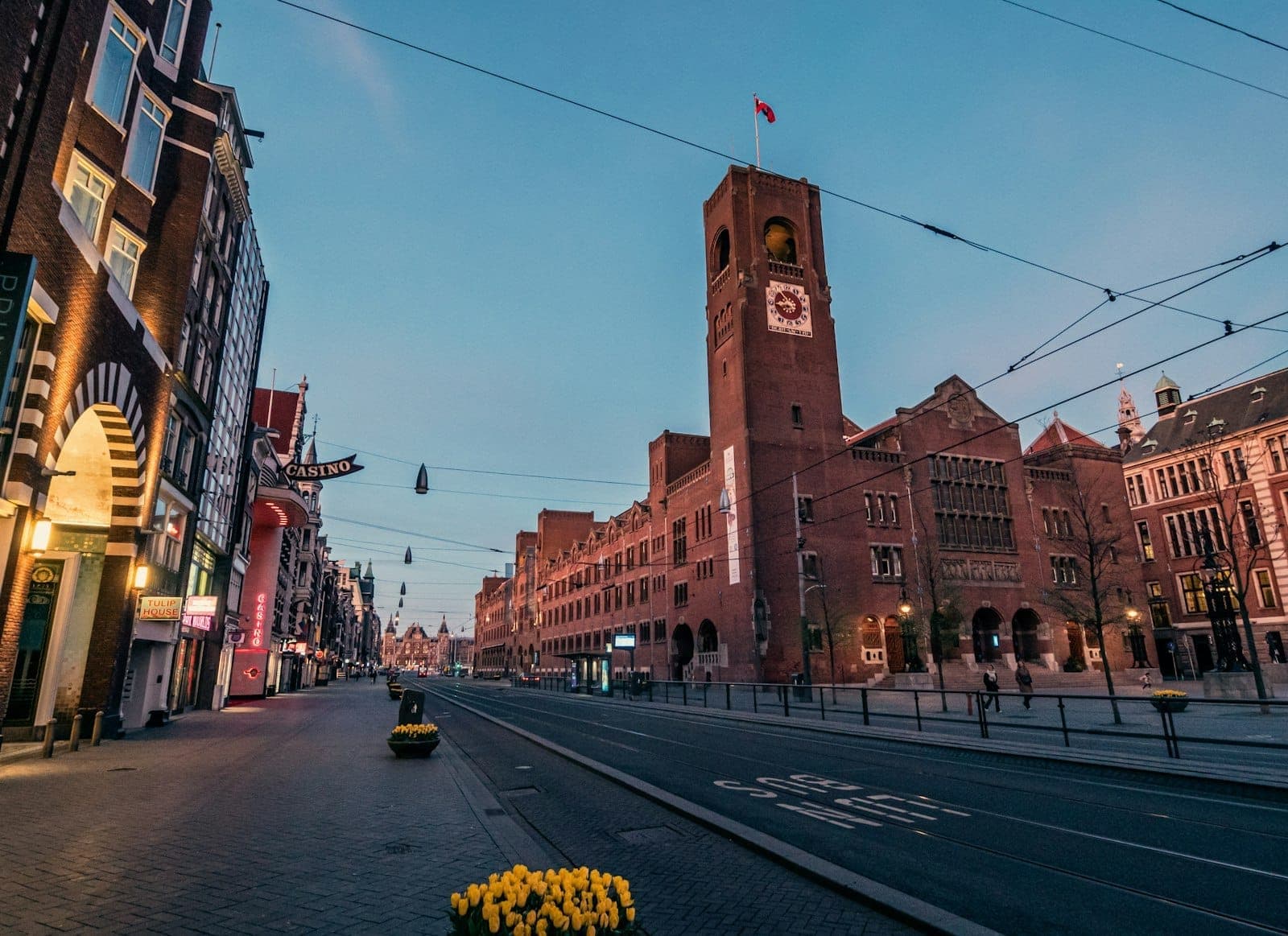 Tampilan sudut lebar fasad bata dan menara jam Beurs van Berlage di jalan Damrak, dengan rel trem dan tulip kuning di latar depan saat senja.