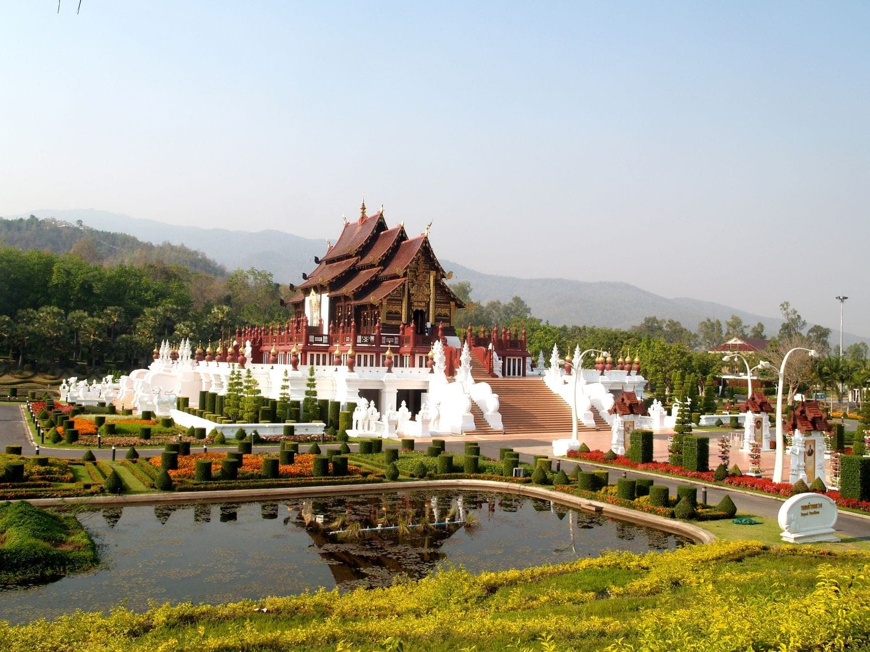 Jardines formales cuidadosamente diseñados y un estanque con un elegante pabellón de estilo tailandés, enmarcado por las laderas boscosas del Doi Suthep en Chiang Mai, bajo un cielo despejado.