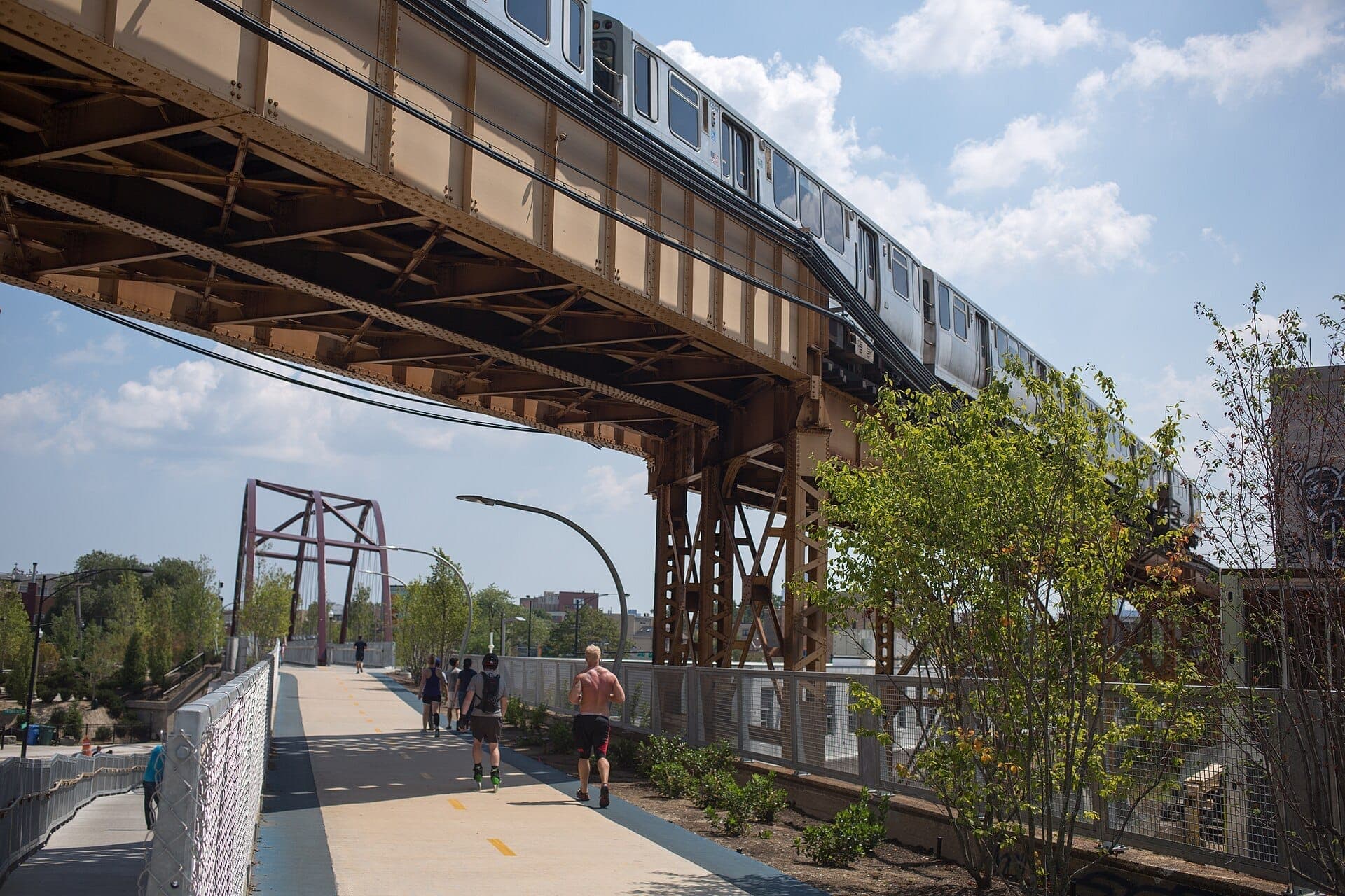 People jogging and walking on the Bloomingdale Trail (The 606) under a passing Chicago elevated train on a sunny afternoon.