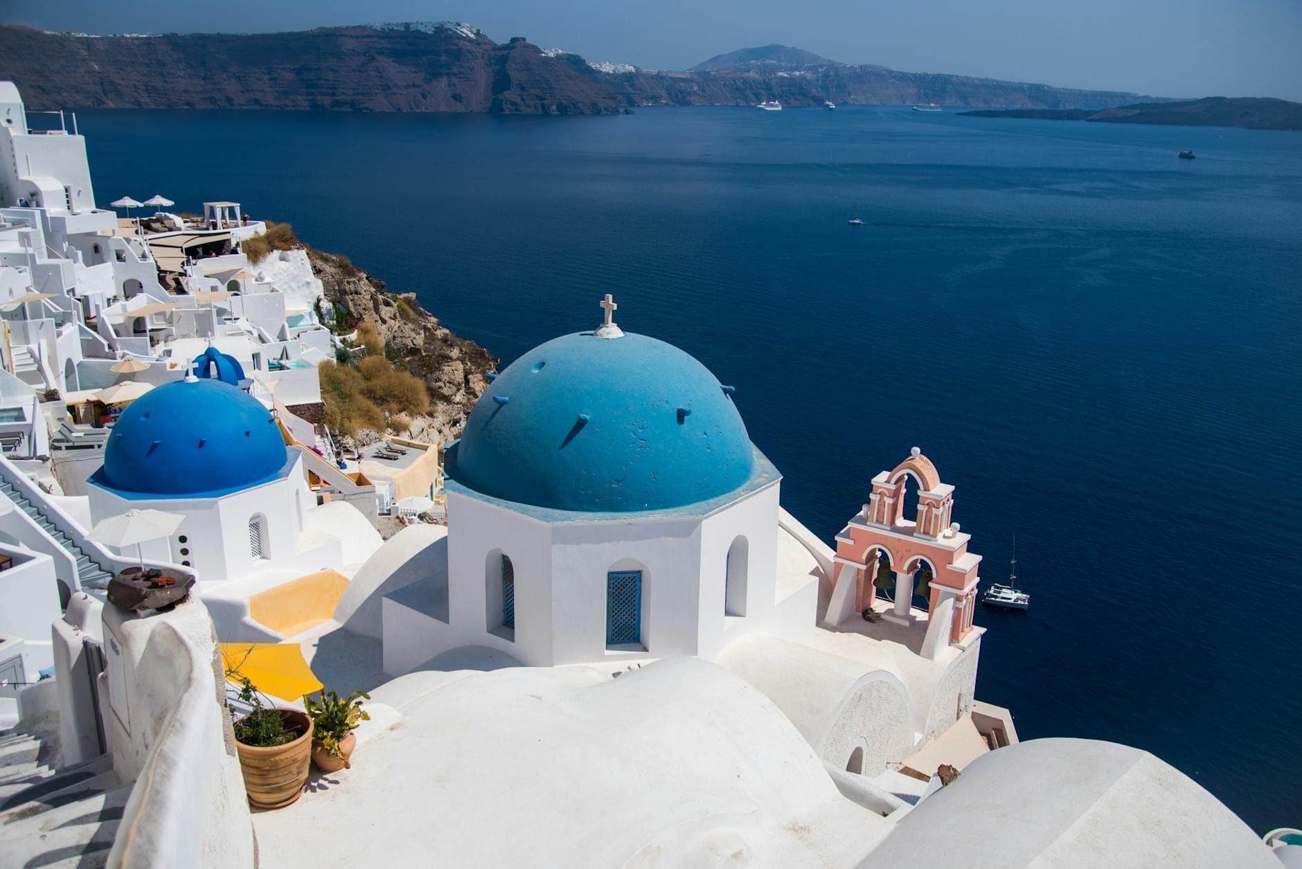 Zwei ikonische Blaukuppelkirchen und ein pfirsichfarbener Glockenturm am Klippenrand über dem tiefblauen Meer in Oia, Santorin, mit weiß getünchten Häusern, die den Hang hinabkaskadieren.