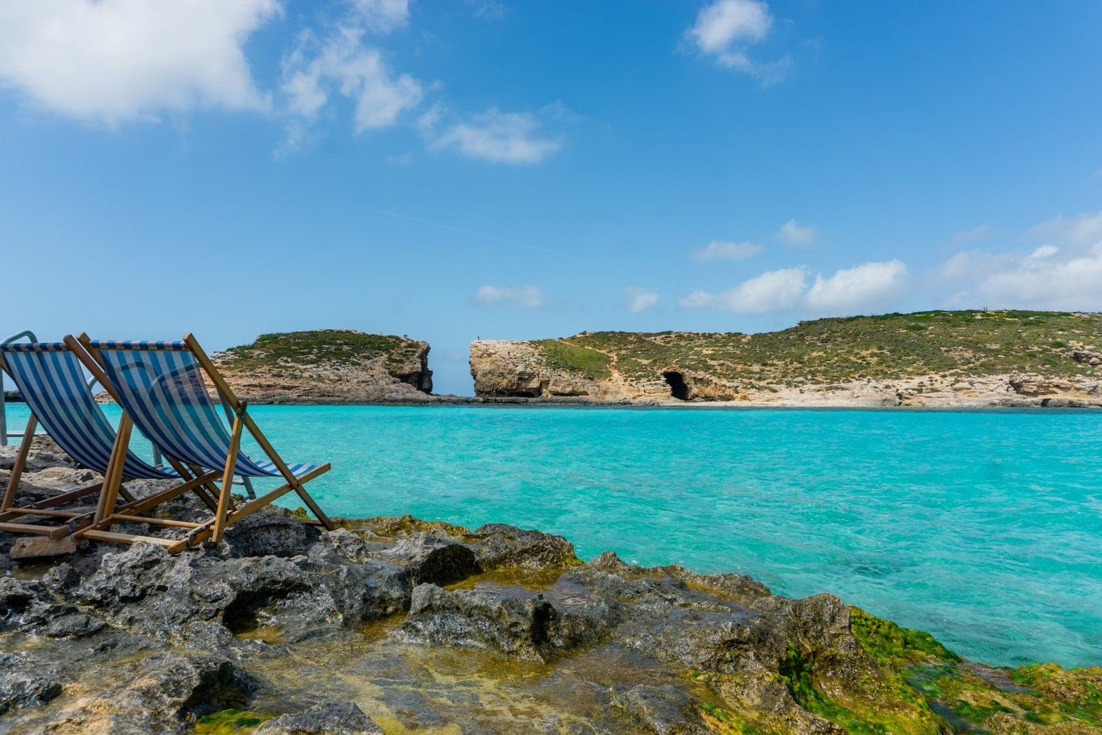 Zwei blau gestreifte Liegestühle an einem felsigen Ufer mit Blick auf das türkisfarbene Wasser und die zerklüfteten Klippen der Blue Lagoon, Comino, unter einem strahlenden Himmel.