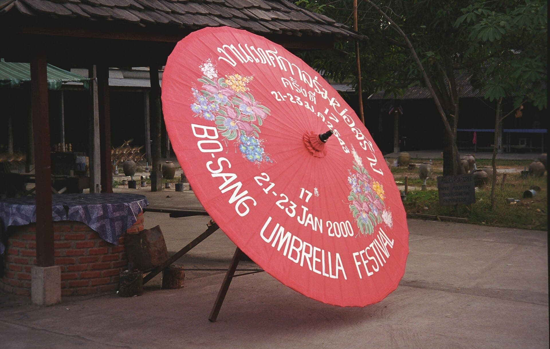 Großer roter Sonnenschirm mit gemalten Blumen und dem Schriftzug des Bo Sang Umbrella Festival, ausgestellt vor einem traditionellen Holzgebäude im Handwerkerdorf.