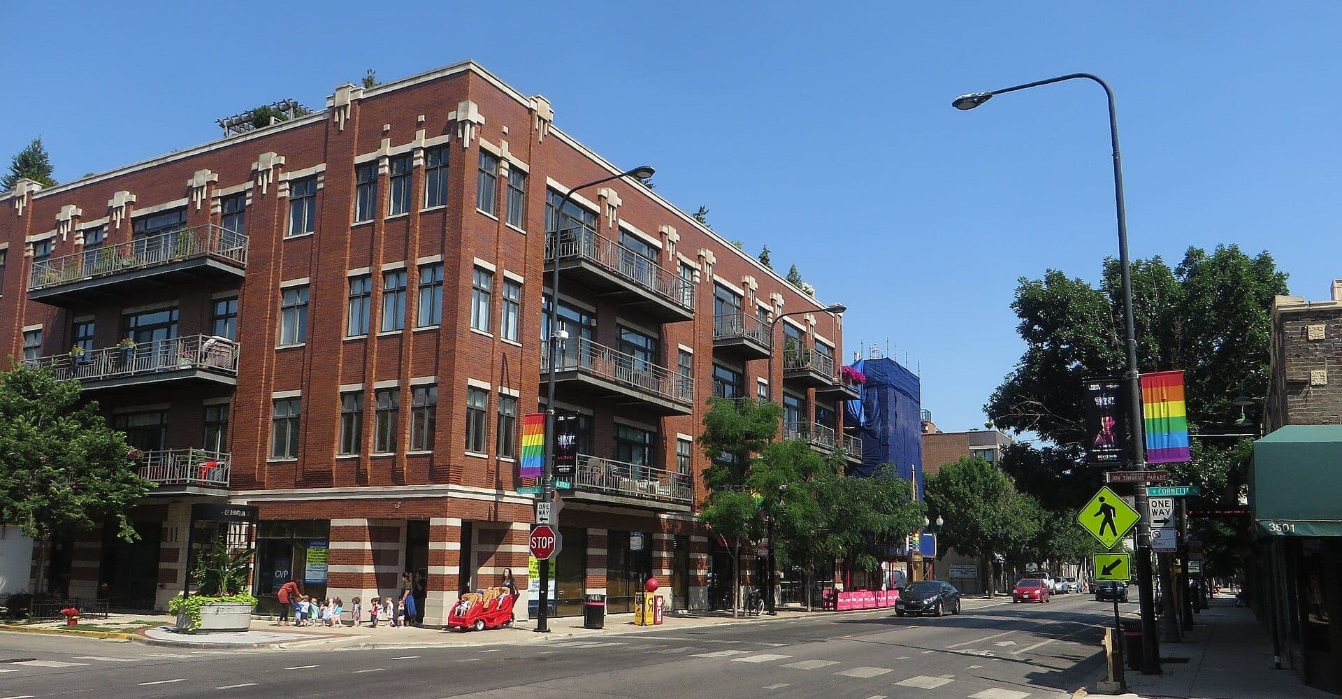 อาคารอพาร์ตเมนต์อิฐแดงบน North Halsted Street ในย่าน Boystown เมืองชิคาโก พร้อมธงไพรด์สีรุ้งและนักเดินเท้าใต้ท้องฟ้าสีฟ้าใส