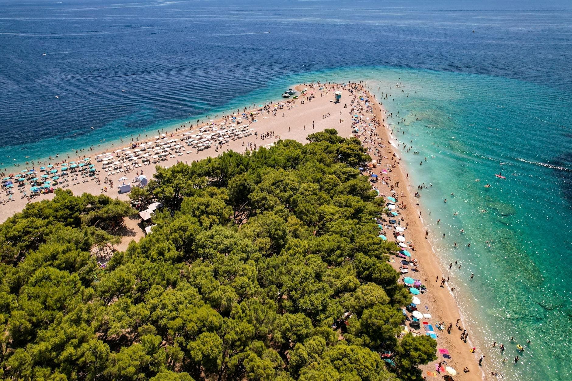 Vista aerea della celebre spiaggia Zlatni Rat sull'isola di Brač, in Croazia, con acque turchesi, bagnanti, ombrelloni e una fitta pineta verde.
