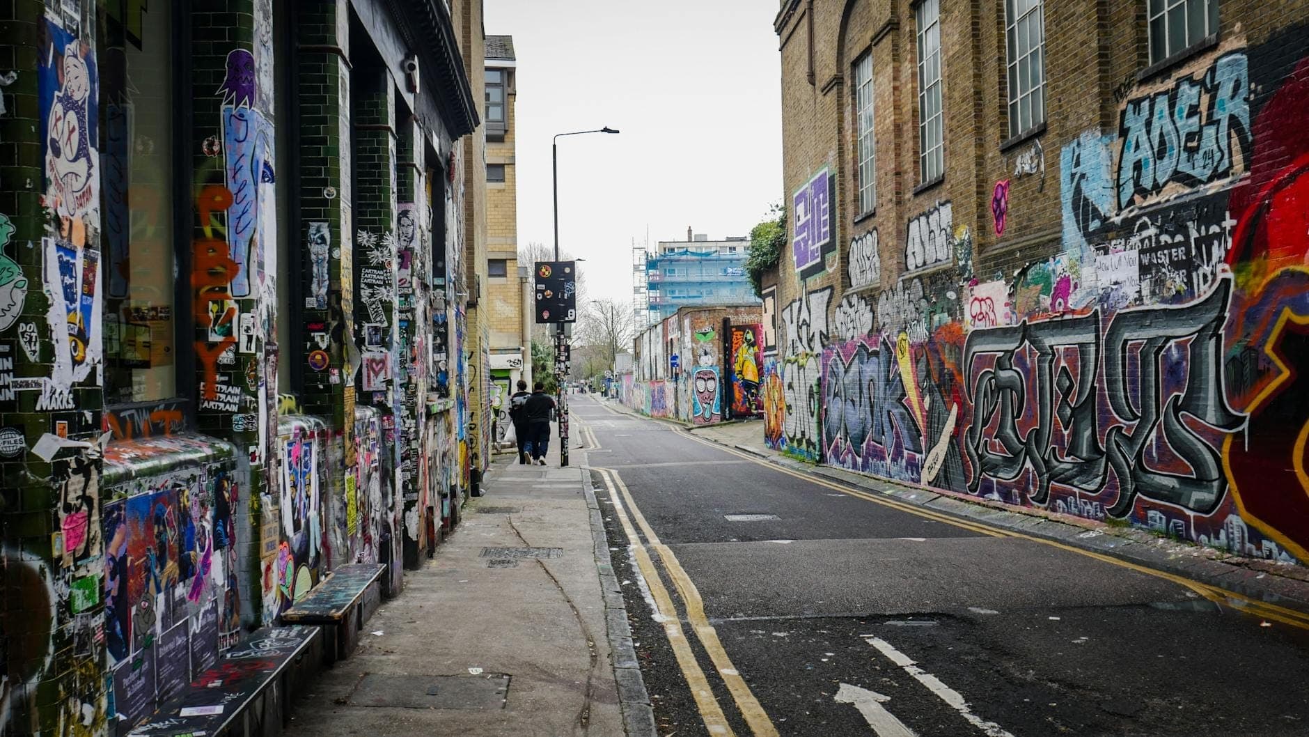 Die bunte Brick Lane mit leuchtendem Graffiti, alten Backsteingebäuden und einigen Passanten in der Ferne an einem bewölkten Tag.