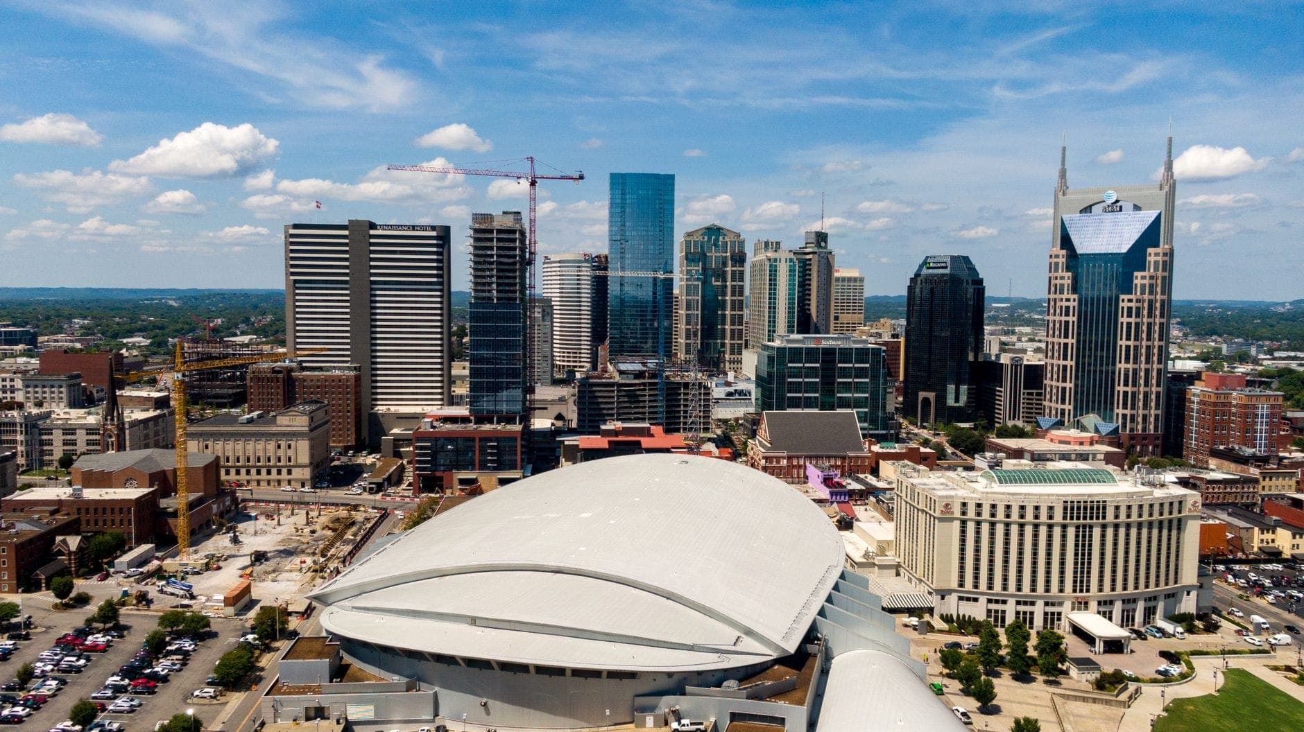 Luftaufnahme der Bridgestone Arena mit ihrem geschwungenen Dach in der Innenstadt von Nashville, umgeben von modernen Wolkenkratzern und Baukränen unter blauem Himmel.