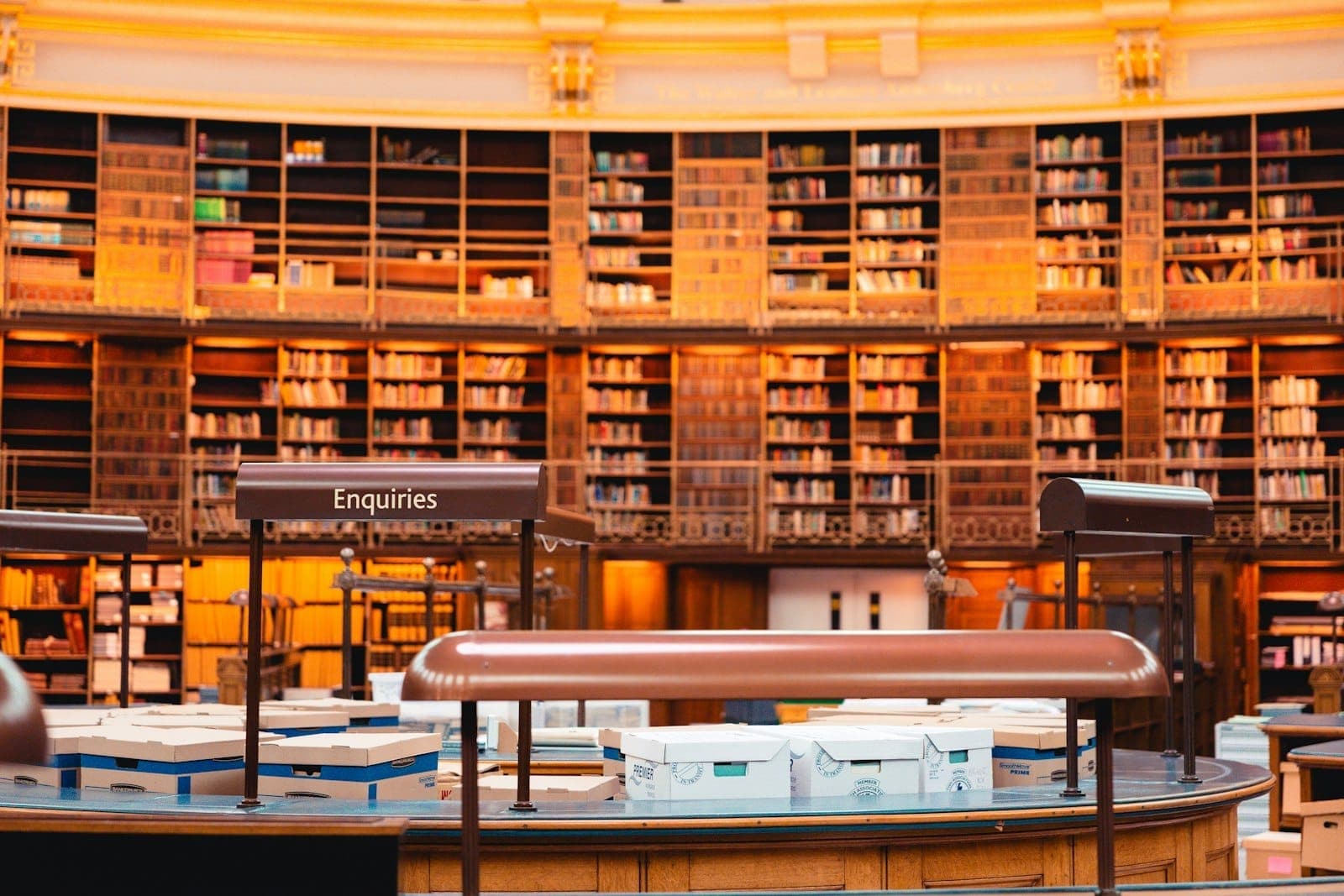 Reihen von Bücherregalen und ein Auskunftsschalter im Inneren des großen Lesesaals der British Library, warm beleuchtet in goldenen Tönen.