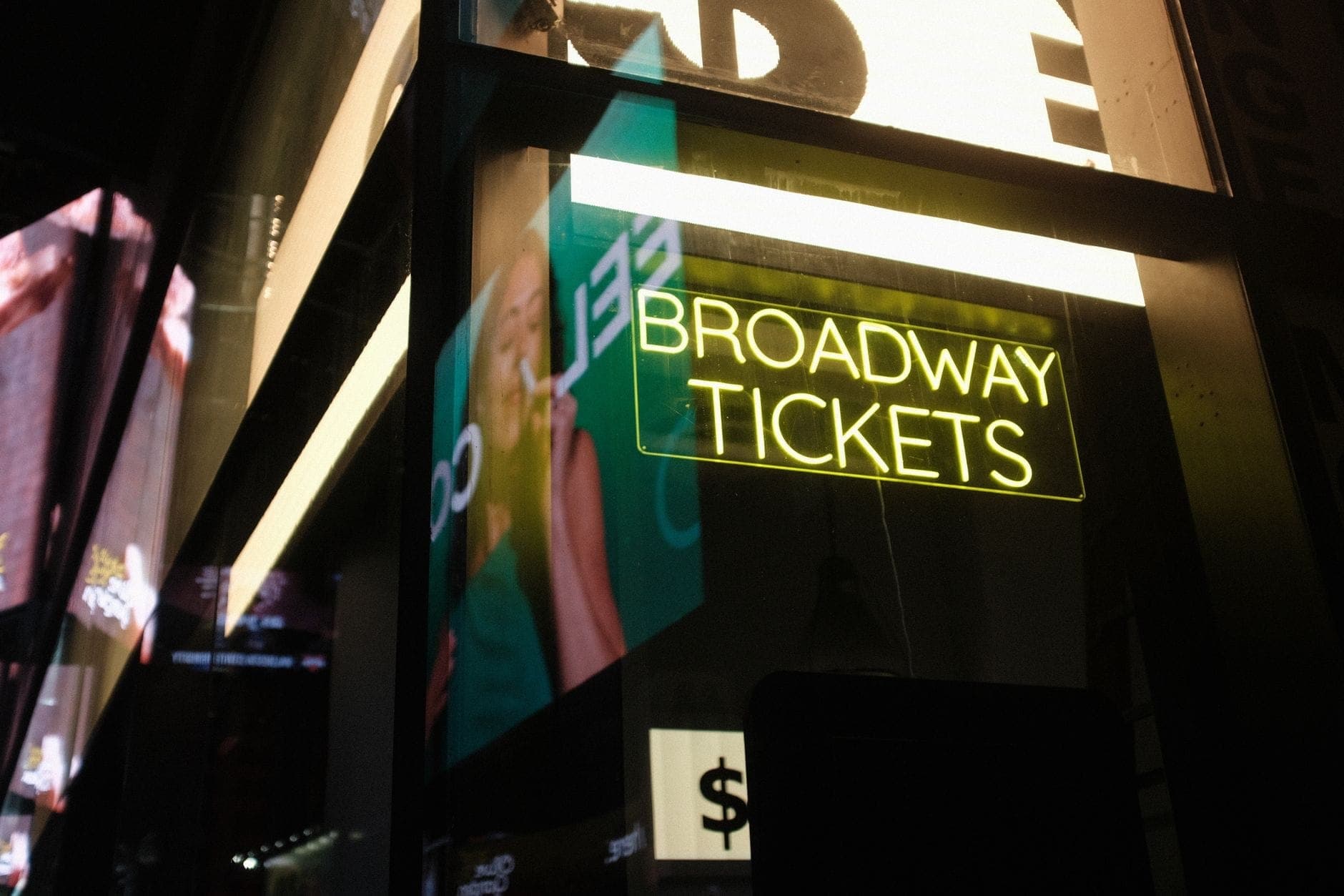 ป้ายนีออนบนหน้าต่างที่มีข้อความว่า 'Broadway Tickets' พร้อมแสงไฟของเมืองและแสงสะท้อน ชวนให้นึกถึงจุดจำหน่ายตั๋วอย่างเป็นทางการใน New York City