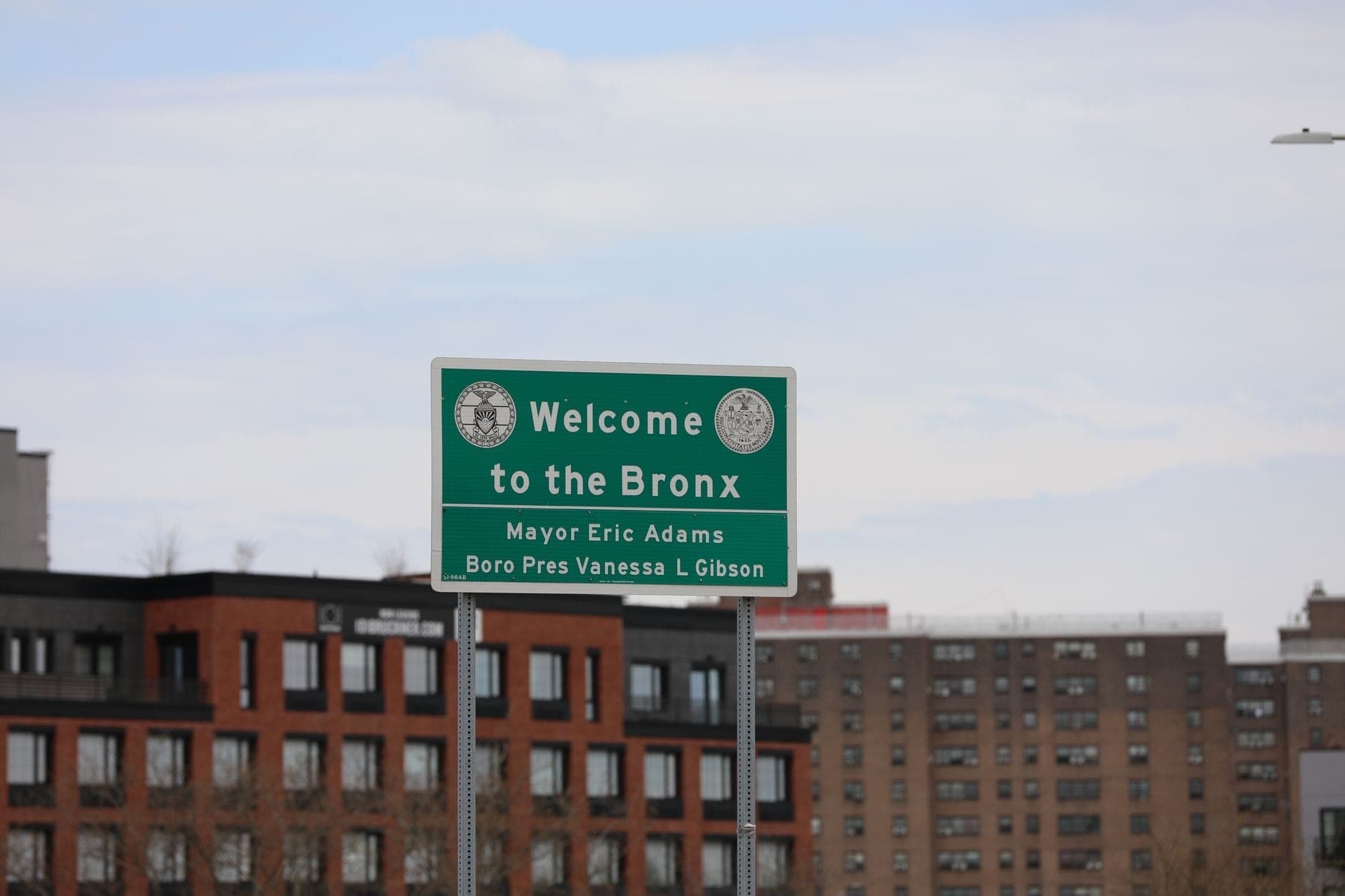 ป้ายสีเขียว 'Welcome to the Bronx' พร้อมอาคารและอพาร์ตเมนต์ในเมืองเป็นฉากหลังใต้ท้องฟ้ามีเมฆบางส่วน
