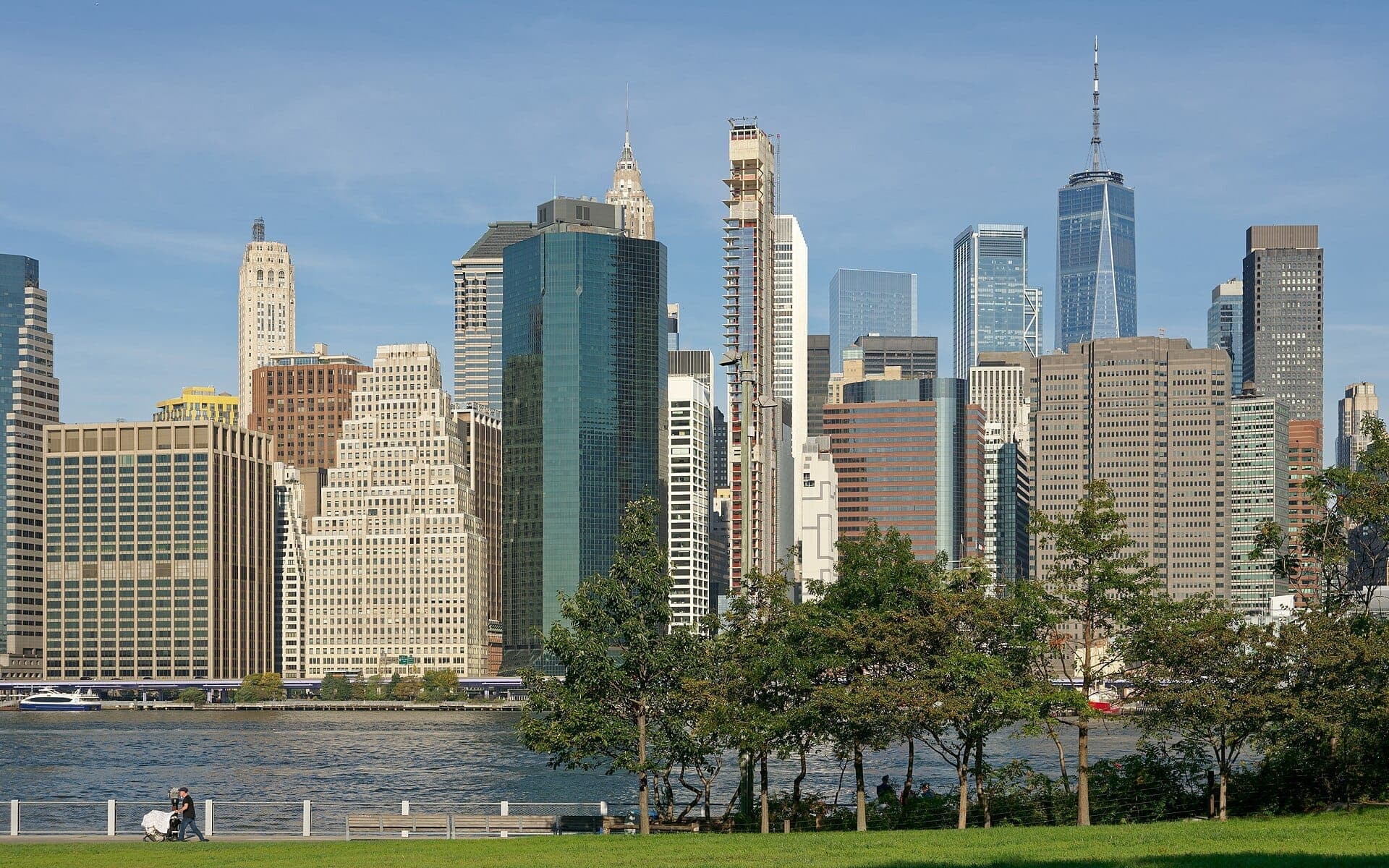 วิวจาก Brooklyn Bridge Park แสดงให้เห็นสนามหญ้าสีเขียว ต้นไม้ และเส้นขอบฟ้าของ Manhattan พร้อมตึกระฟ้าสูงเหนือฝั่ง East River ใต้ท้องฟ้าสีฟ้า