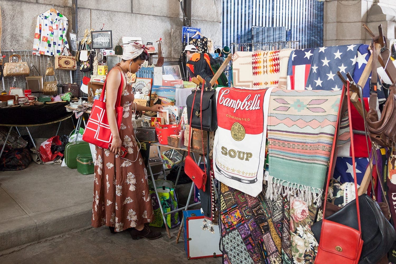 ผู้หญิงคนหนึ่งกำลังเลือกดูกระเป๋าวินเทจและสิ่งทอที่ Brooklyn Flea DUMBO พร้อมด้วยผ้าห่มหลากสี ของตกแต่งสไตล์เรโทร และแผงตลาดใต้ซุ้มโค้งในเมือง