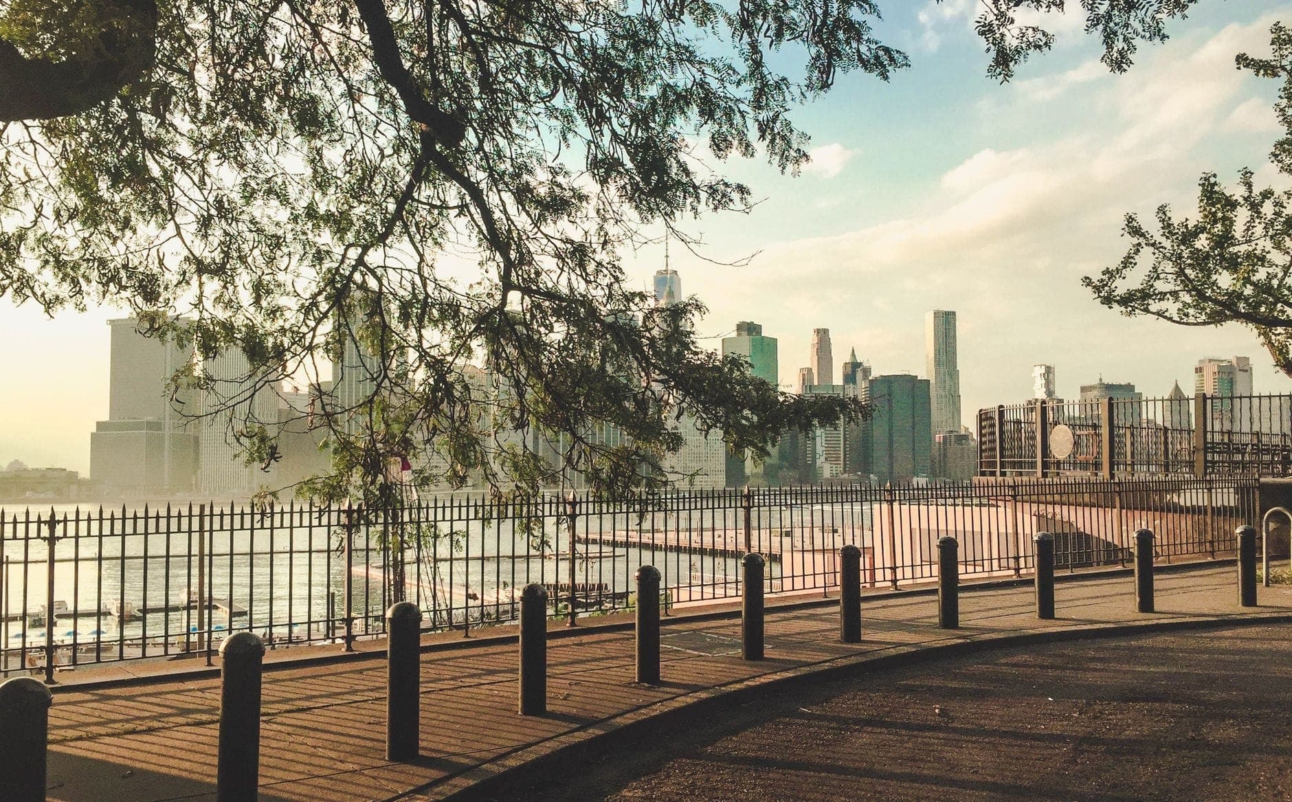 วิวจาก Brooklyn Heights Promenade มองเห็นแมนฮัตตัน New York Harbor และ Brooklyn Bridge ต้นไม้เรียงรายส่องแสงแดดริมทางเดินชมวิวริมแม่น้ำ