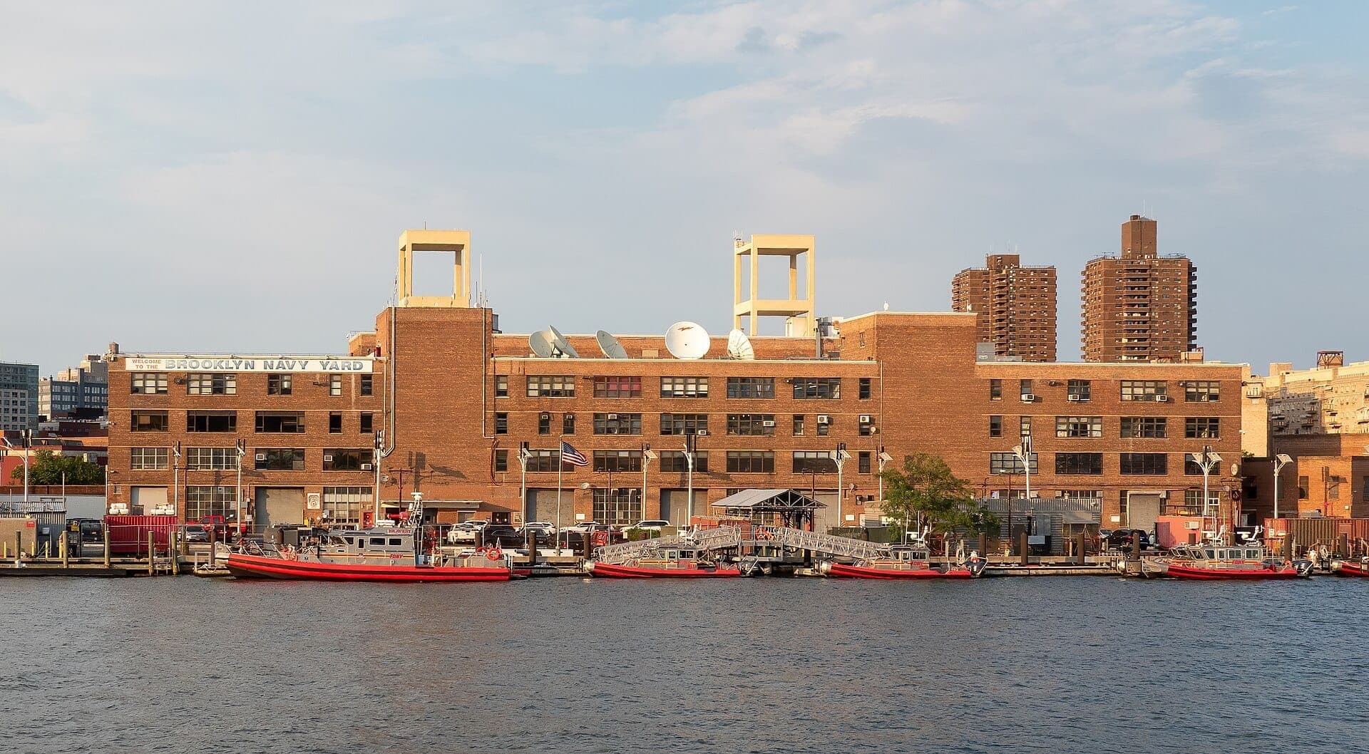 ท่าเรืออุตสาหกรรม Brooklyn Navy Yard พร้อมอาคารอิฐ จานดาวเทียม เรือสีแดงจอดเทียบท่า และตึกระฟ้าของเมืองในพื้นหลังในวันที่มีแดดจ้า