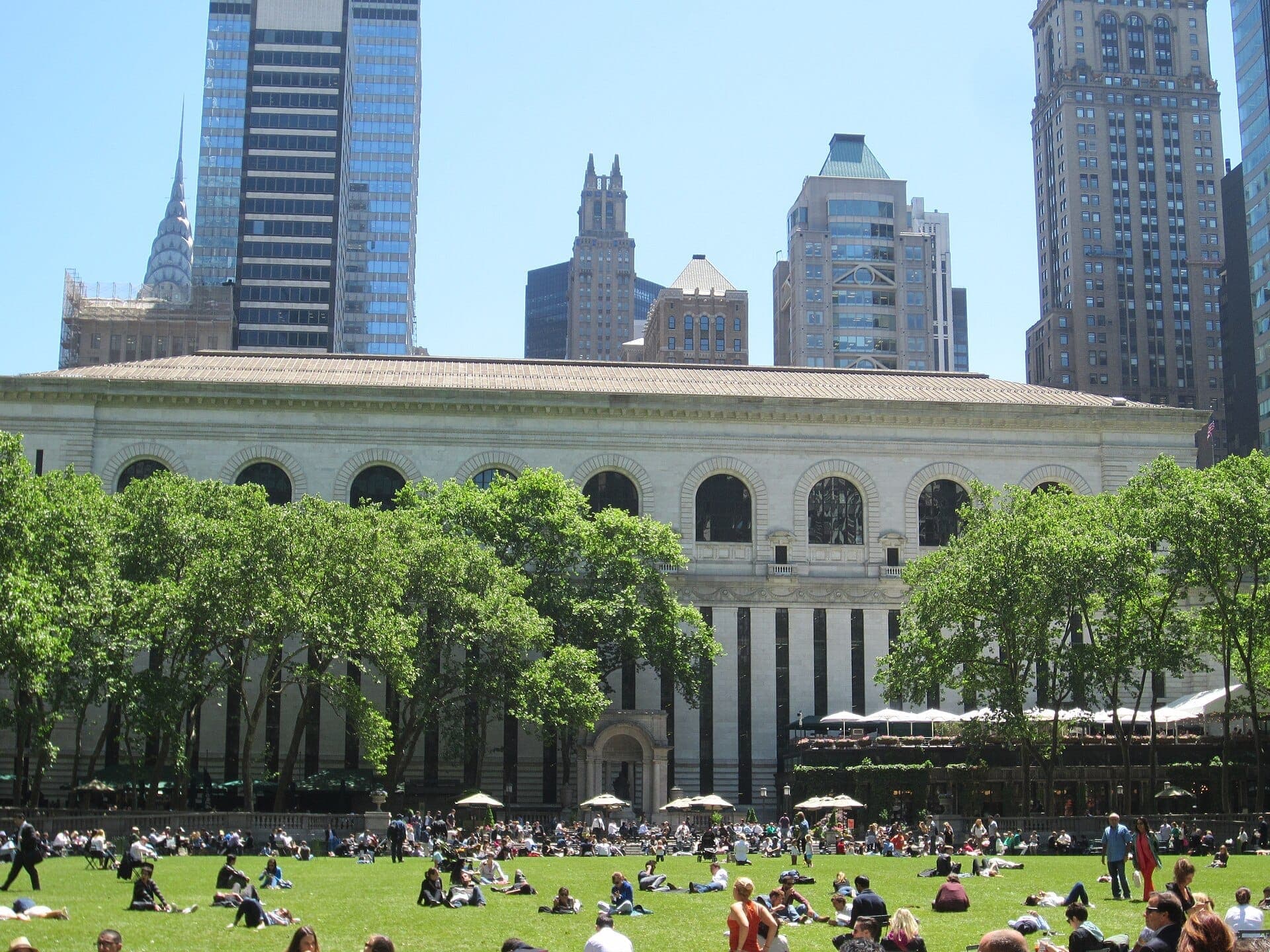 Bryant Park ในวันที่แดดจ้า มีผู้คนพักผ่อนบนสนามหญ้า ล้อมรอบด้วยต้นไม้สีเขียวและตึกระฟ้าสัญลักษณ์ของ Midtown Manhattan