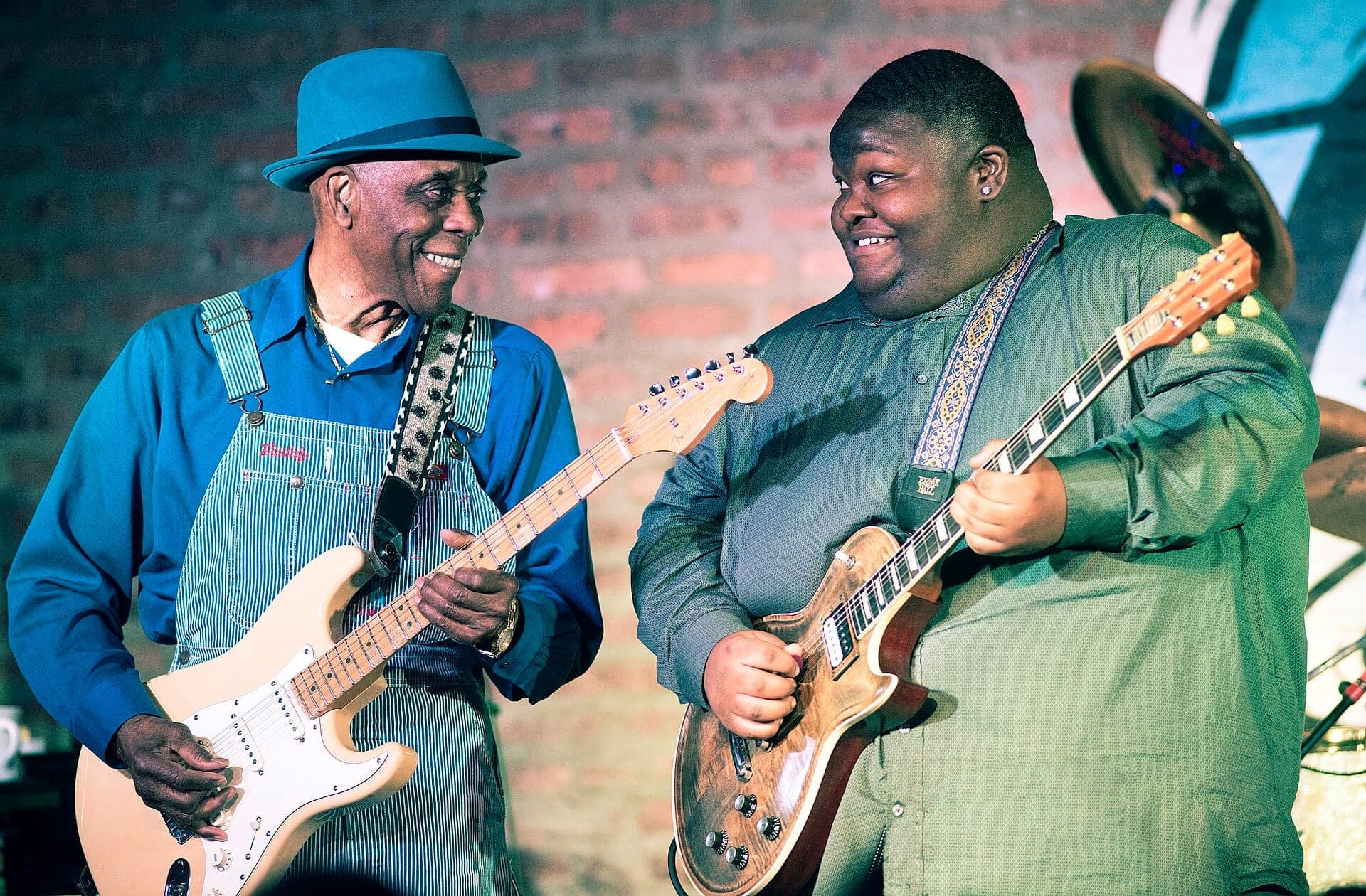 นักดนตรีสองคนเล่นกีตาร์ไฟฟ้าบนเวที Buddy Guy's Legends โดยมีฉากหลังเป็นกำแพงอิฐและแสงไฟเวทีสว่างจ้า