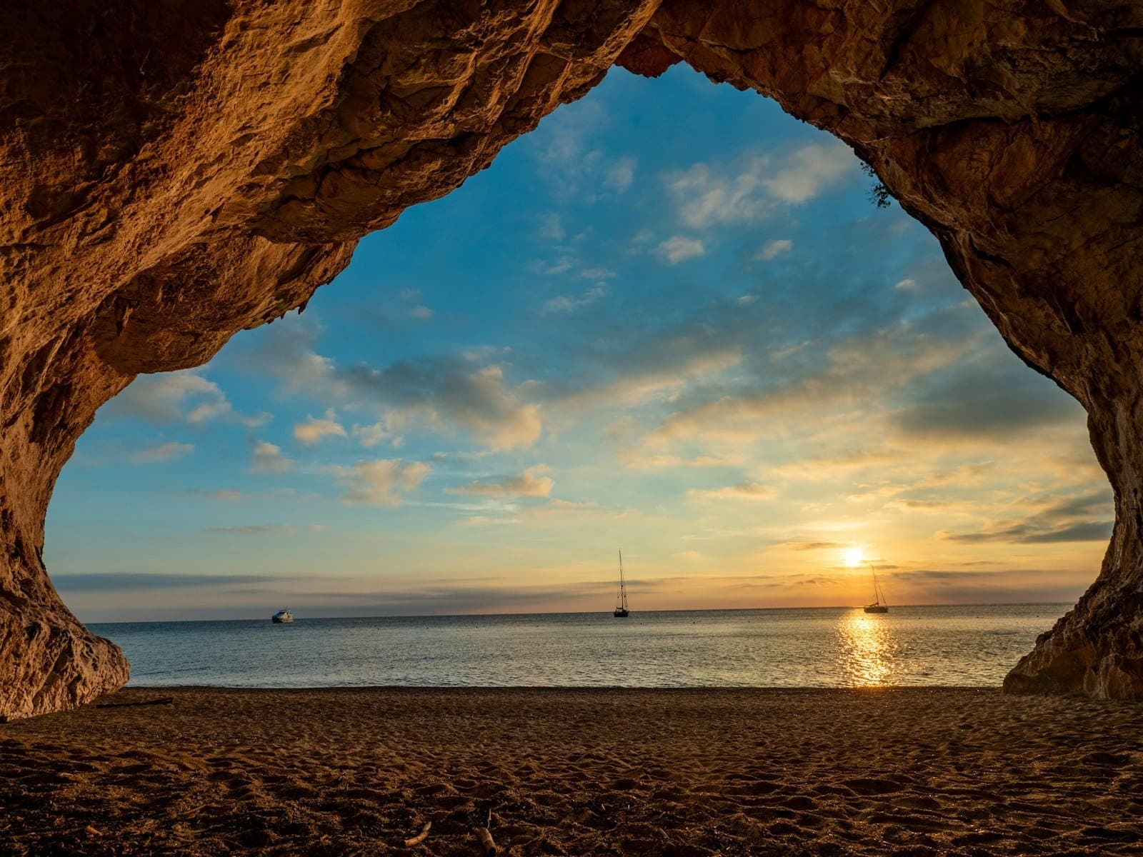 Blick aus der Felshöhle von Cala Luna auf den Sandstrand, das ruhige Meer und Segelboote bei Sonnenuntergang auf Sardinien, Italien.