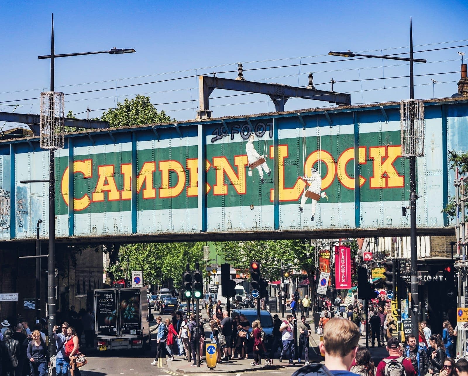 Scène de rue à Camden sous le célèbre pont ferroviaire de Camden Lock avec ses lettres jaunes vives, une foule animée et un ciel bleu au-dessus.