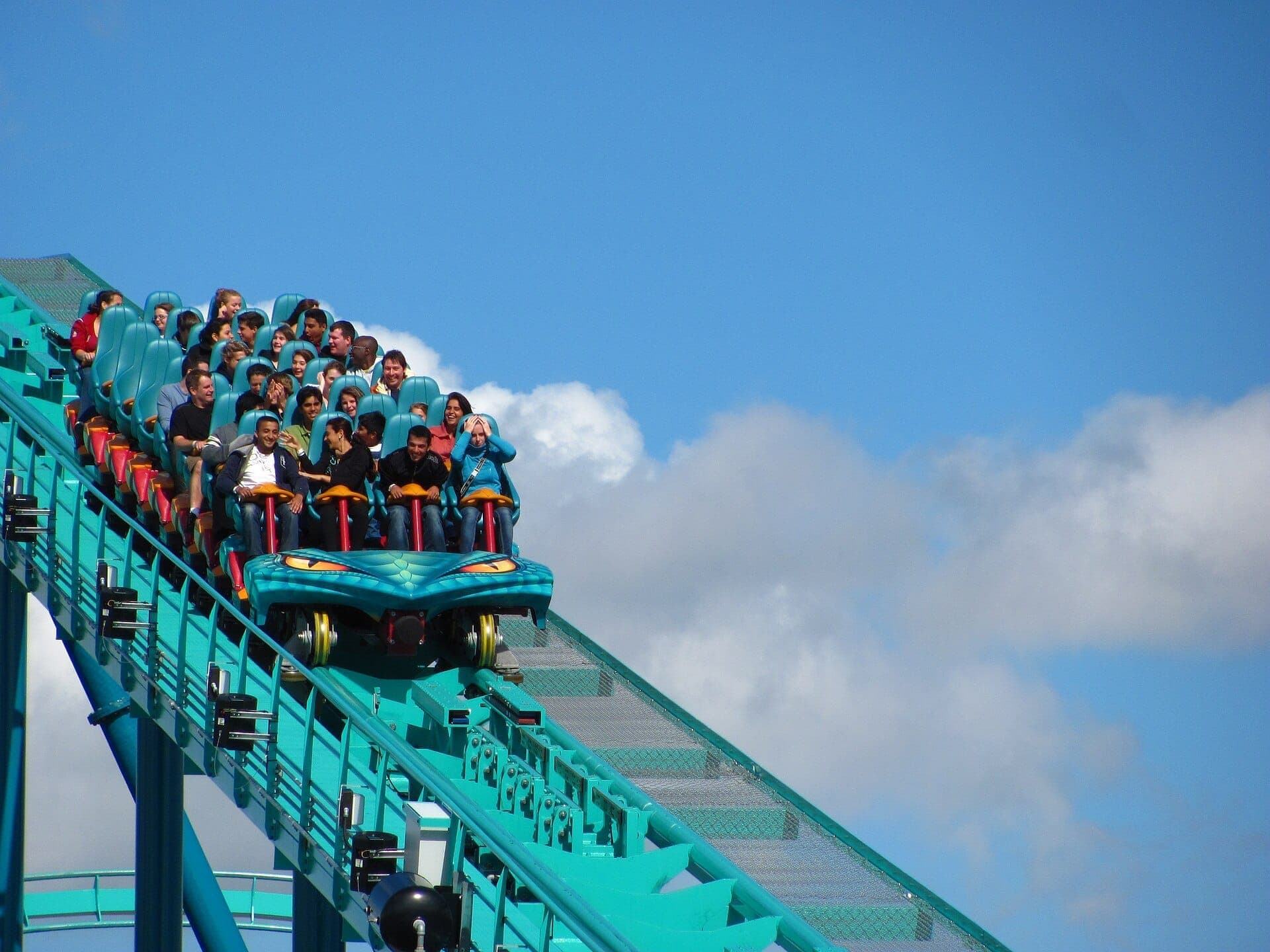 Un vibrante vagón de montaña rusa lleno de emocionados pasajeros sube por una vía de color verde azulado bajo un cielo azul brillante en Canada's Wonderland.