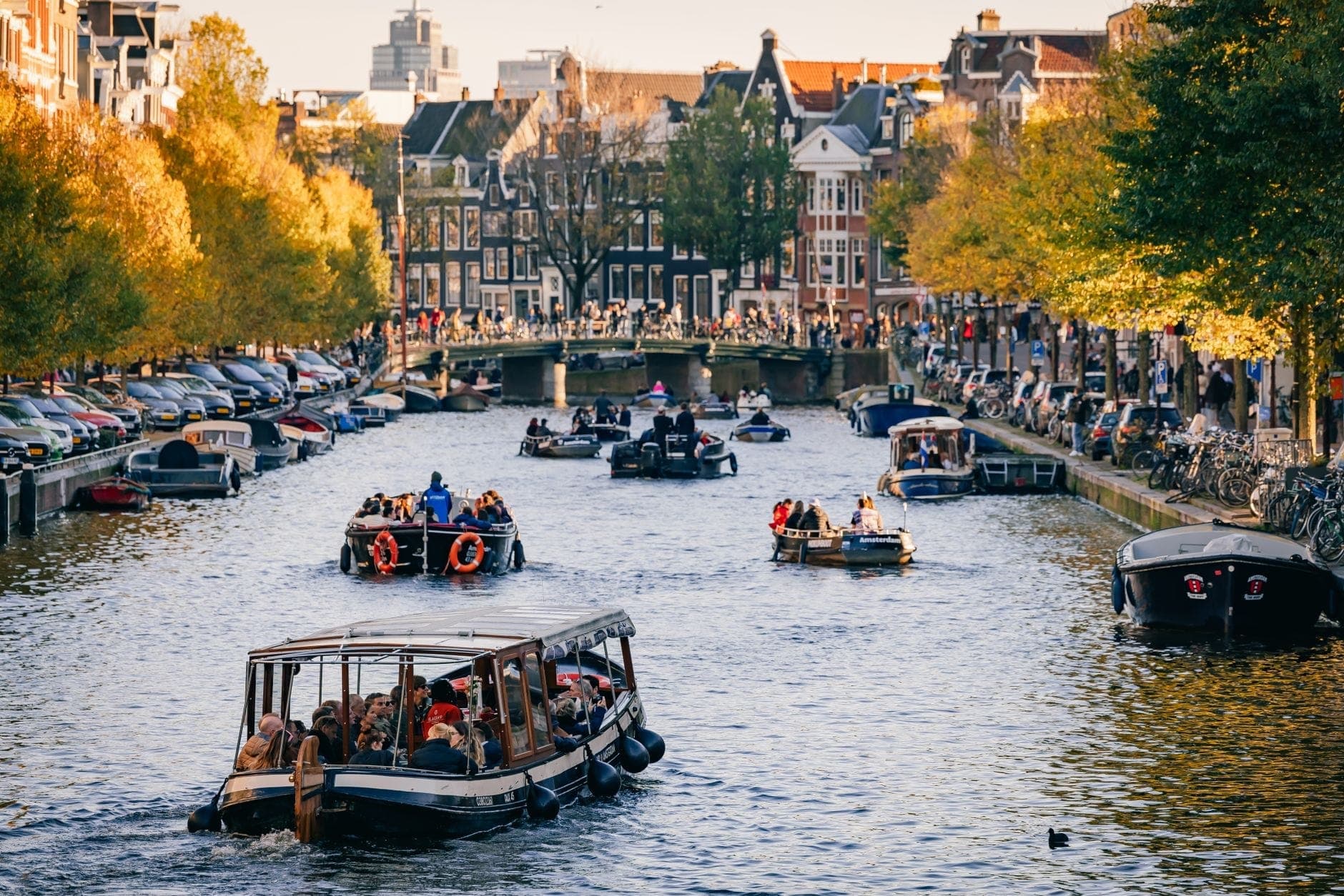 Mehrere Ausflugsboote mit Fahrgästen fahren an einem sonnigen Nachmittag durch einen Amsterdamer Kanal, gesäumt von Bäumen und historischen Gebäuden.