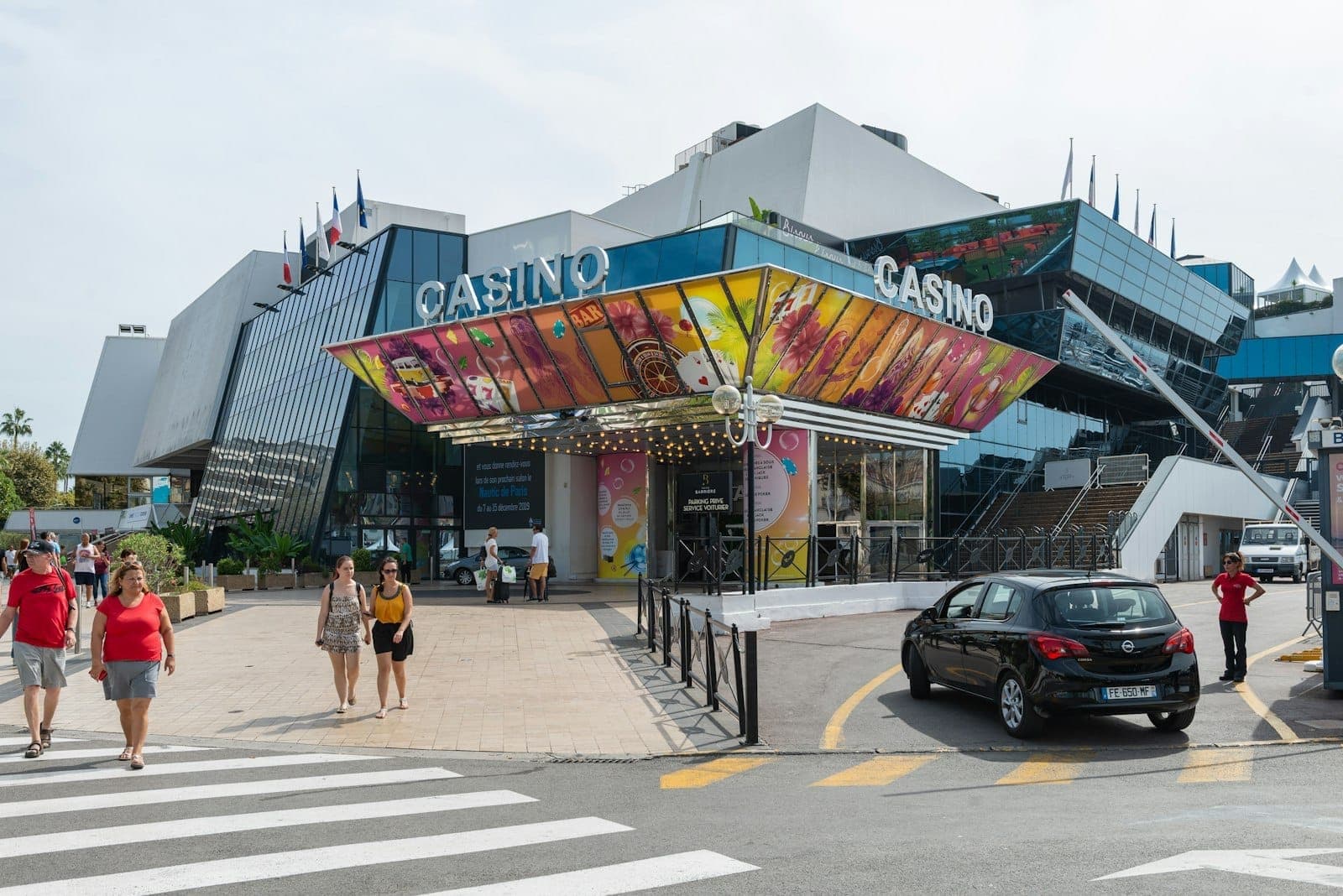 Современное стеклянное здание Palais des Festivals et des Congrès, место проведения Каннского кинофестиваля, с людьми, идущими снаружи, и припаркованными перед ним автомобилями.