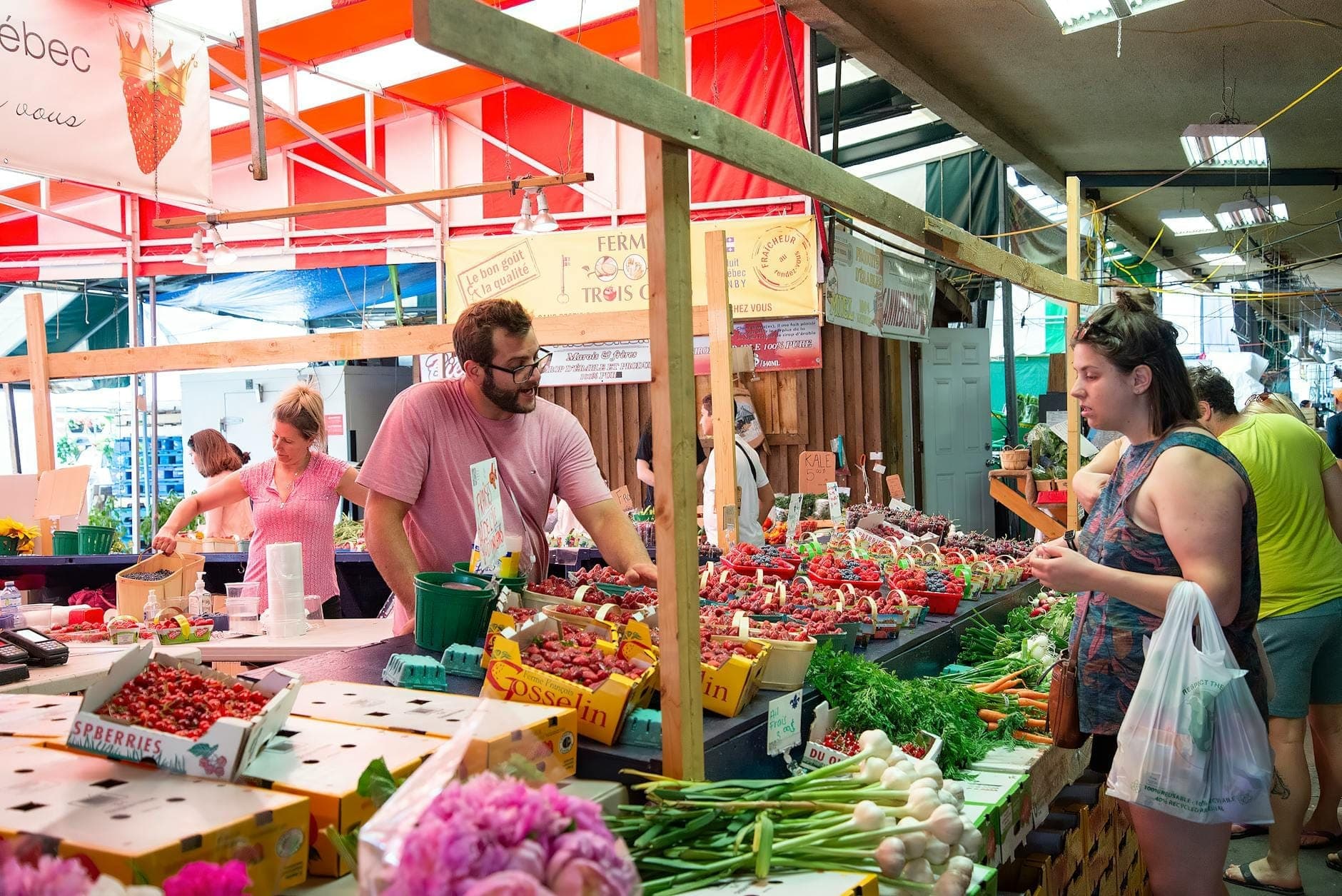 Сцена крытого рынка с продавцами, предлагающими покупателям свежие продукты, включая клубнику и овощи, под разноцветными навесами.