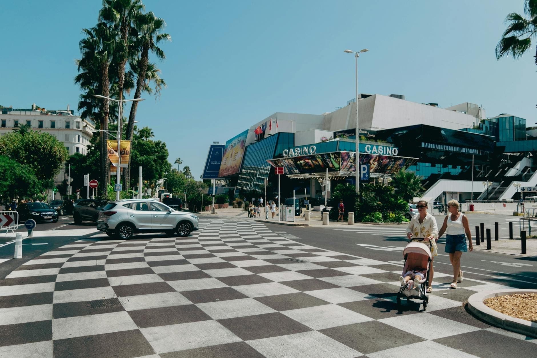 Пешеходы прогуливаются рядом с Palais des Festivals в центре Канн среди пальм, пешеходного перехода и городских зданий в солнечный день.