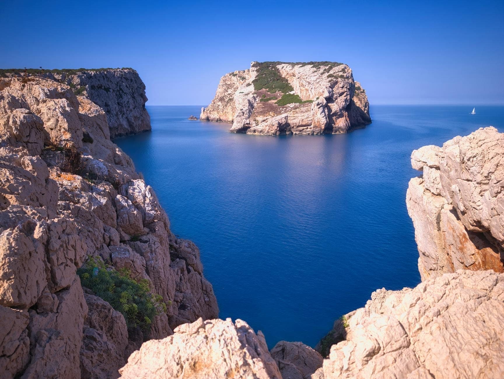 Los imponentes acantilados de caliza y el abrupto promontorio de Capo Caccia se elevan sobre un mar de color azul intenso, vistos desde un mirador rocoso bajo un cielo despejado.