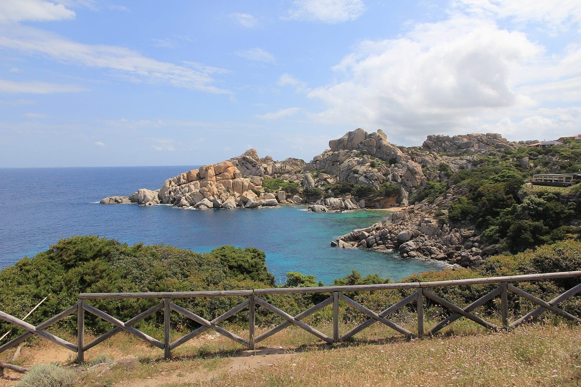 Granitfelsen und üppiges Grün mit Blick auf das klare blaue Meer bei Capo Testa, Sardinien, unter einem teilweise bewölkten Himmel.