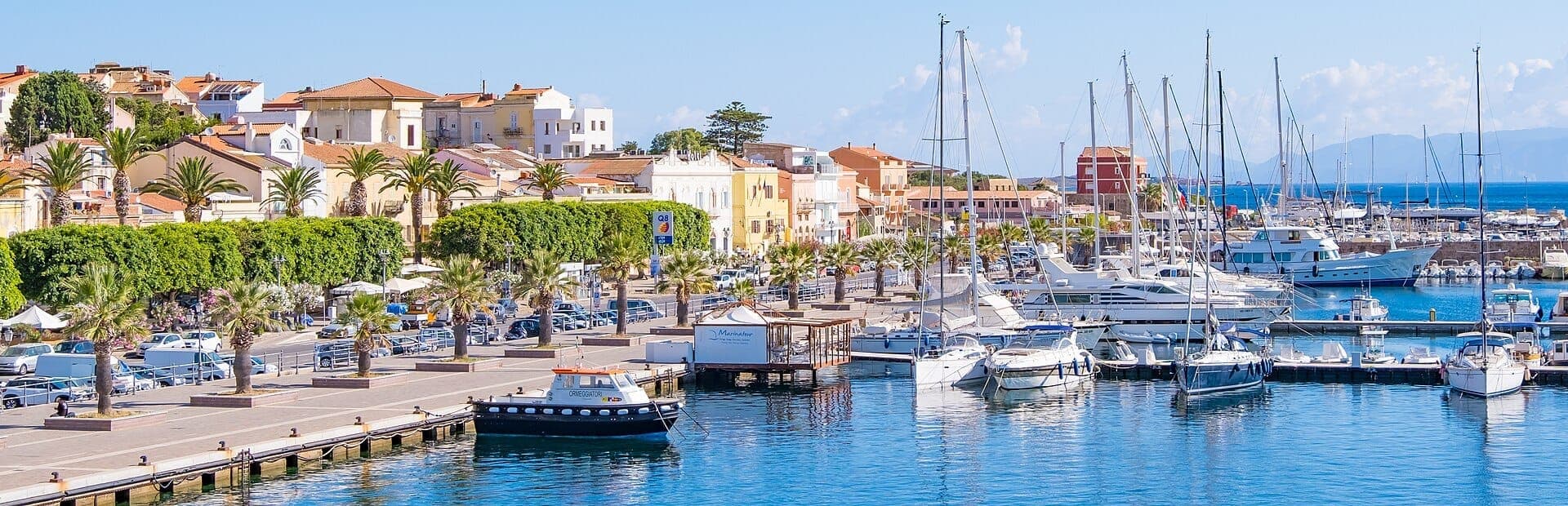 Coloridos edificios bordean el paseo marítimo de Carloforte, con palmeras y veleros atracados que se reflejan en las tranquilas aguas azules del puerto.