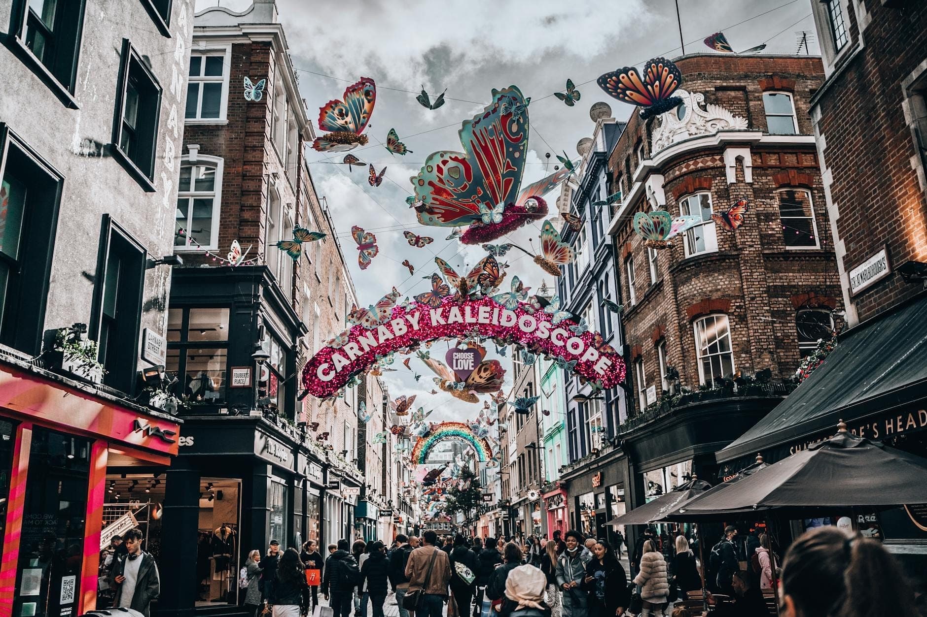 Londra'daki kalabalık Carnaby Street; renkli dükkan cepheleri, asılı kelebek süslemeleri ve 'Carnaby Kaleidoscope' yazan şenlikli bir kemer ile insanlarla dolu.