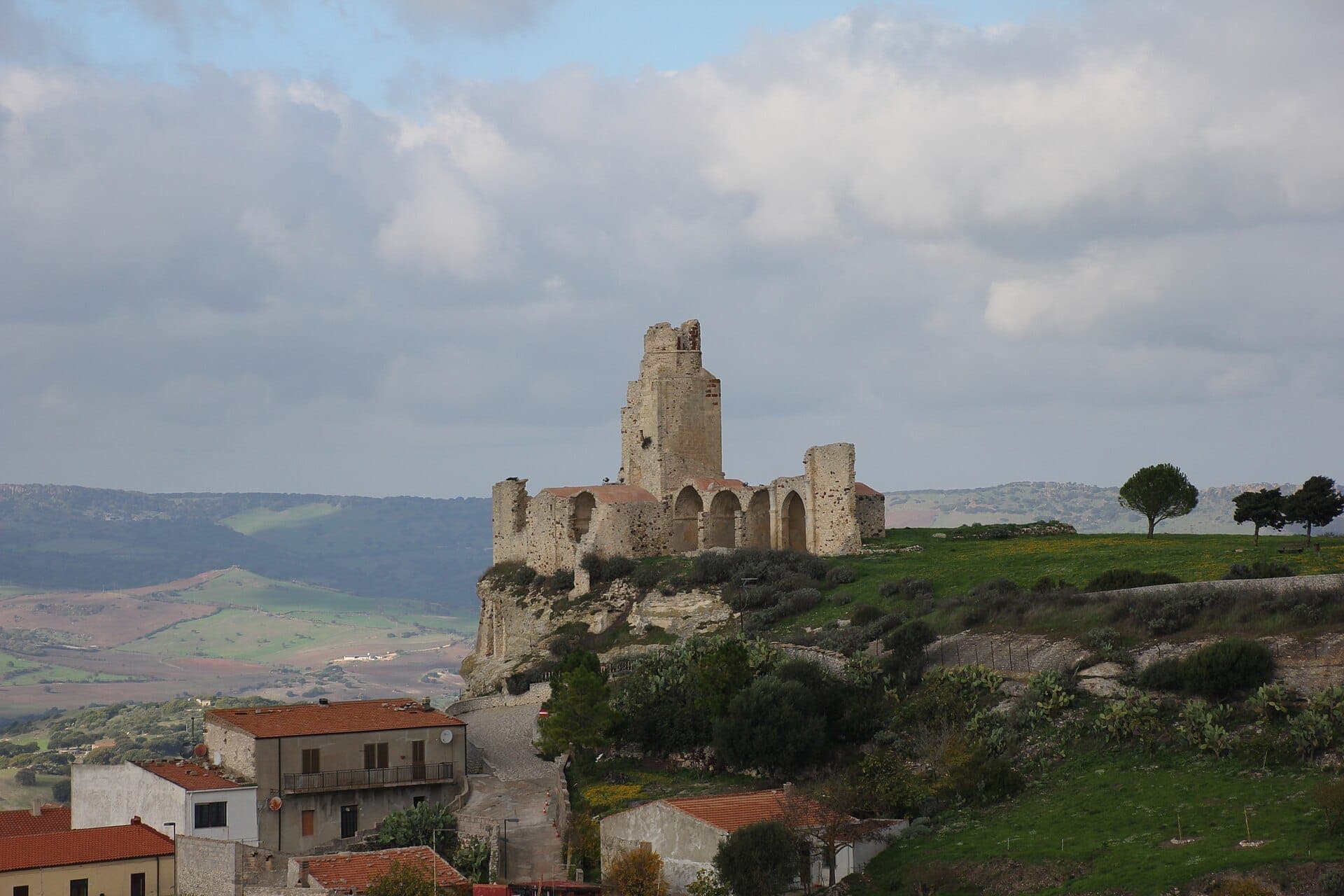Las ruinas de piedra del Castello dei Doria se alzan en una colina verde con vistas a casas y colinas onduladas bajo un cielo nublado en Castelsardo.