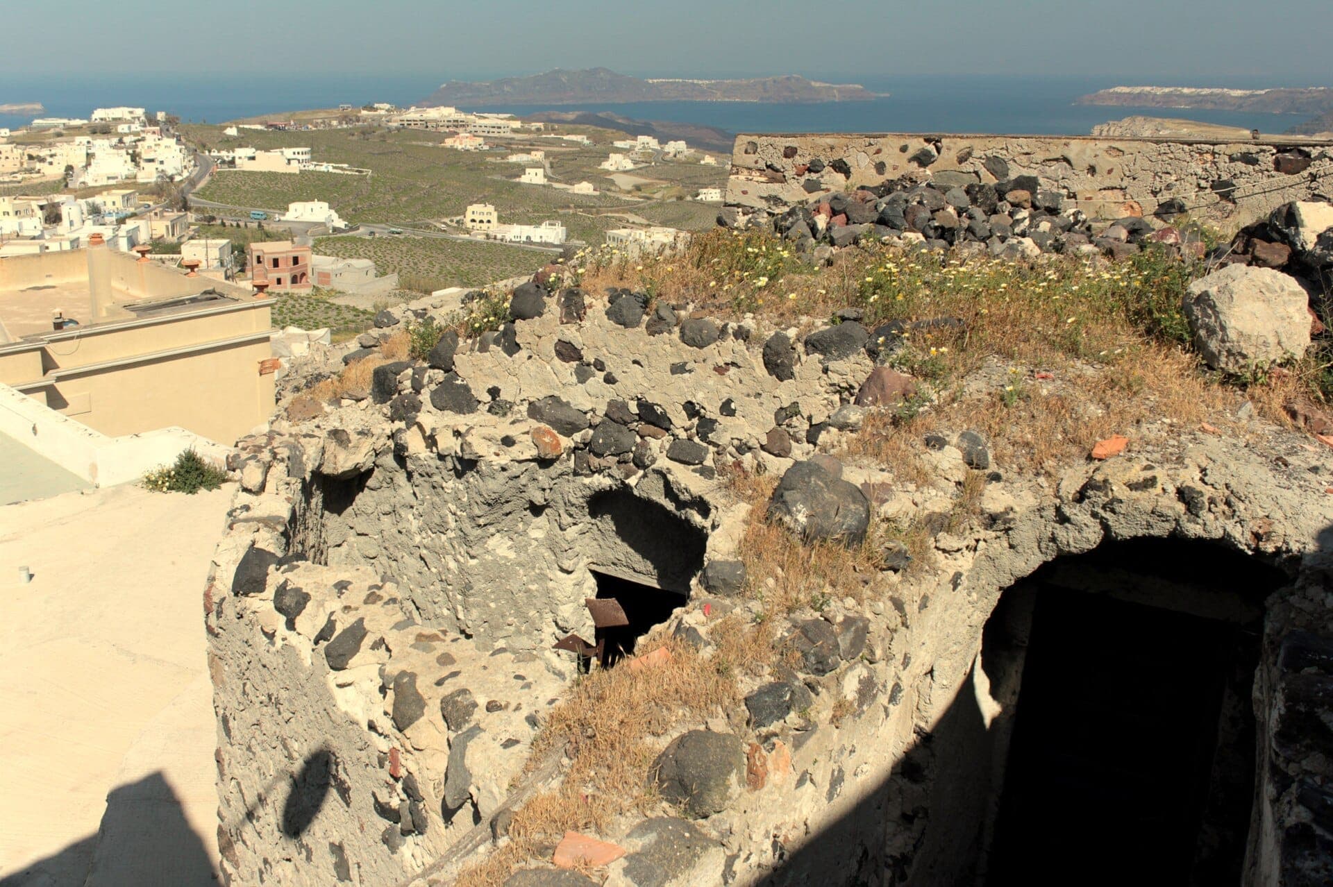 Steinruinen des Schlosses Pyrgos mit Wildblumen, mit Blick auf weiß getünchte Häuser, grüne Felder und das blaue Ägäische Meer in Santorini.