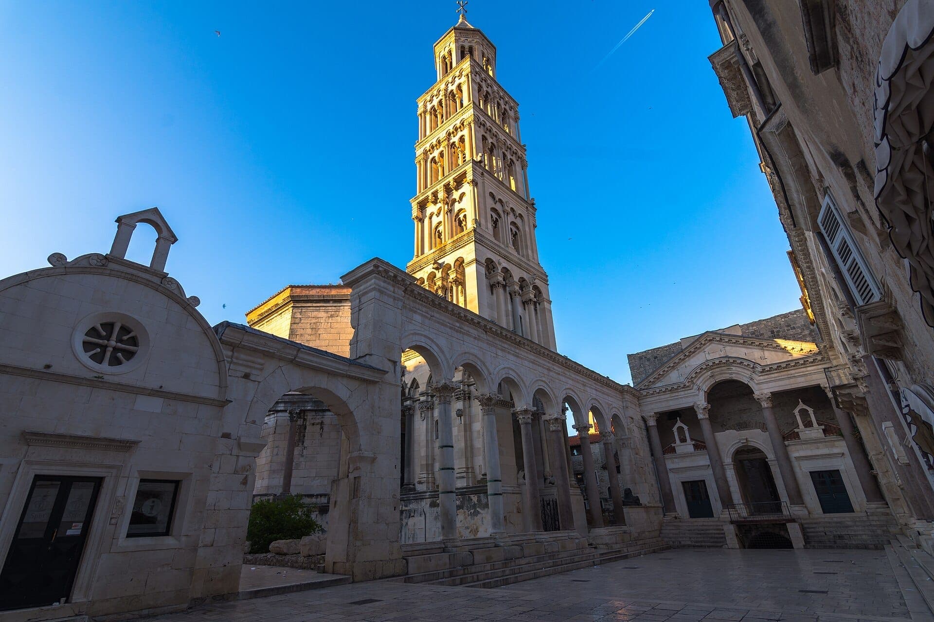 Il campanile della Cattedrale di San Domnius si innalza sopra antichi archi in pietra e piazze, illuminato dalla luce del primo mattino contro un cielo azzurro limpido.
