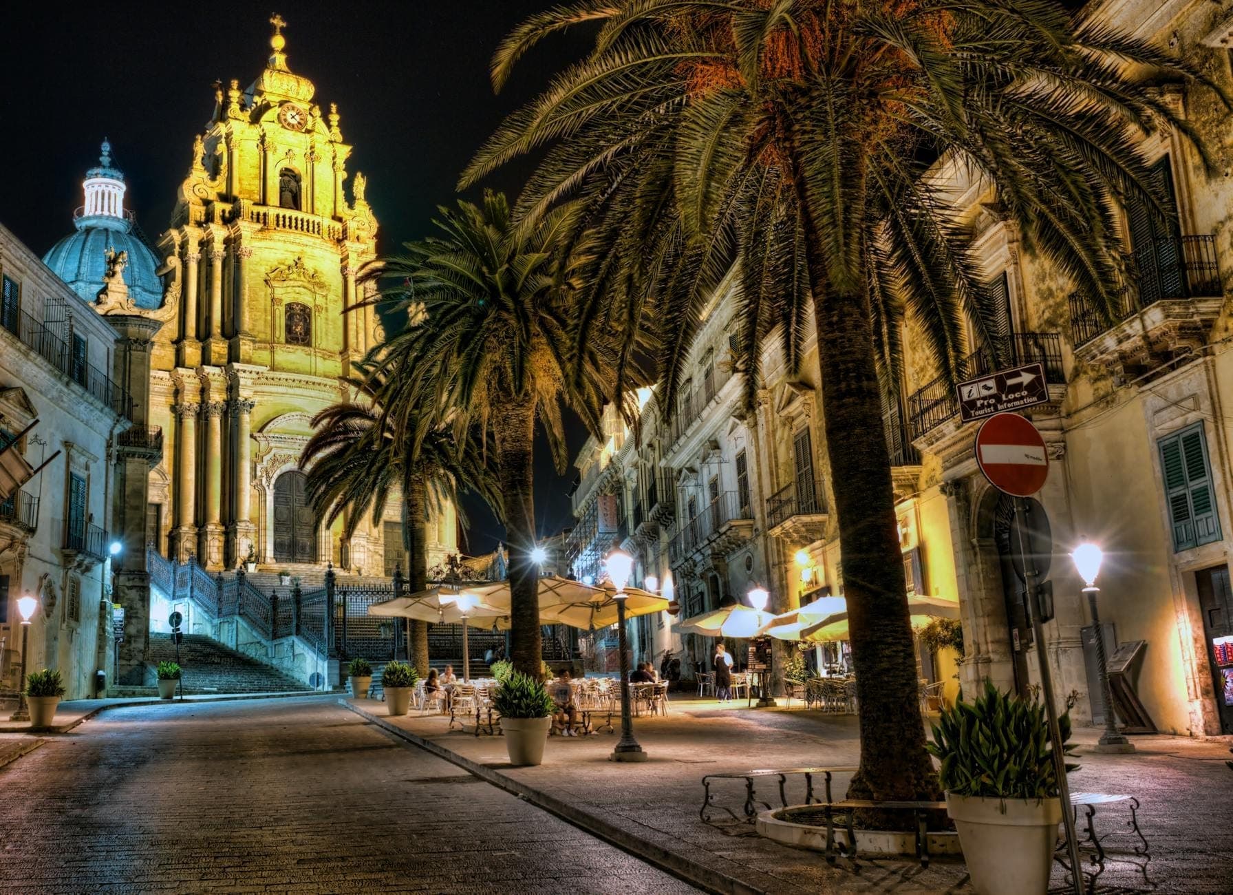 Pemandangan sore Katedral San Giorgio di Ragusa Ibla, menyala dramatis dengan fasad baroknya dan kubah yang menjulang di atas Piazza Duomo yang dihiasi pohon palem dan kafe-kafe di sekitarnya.