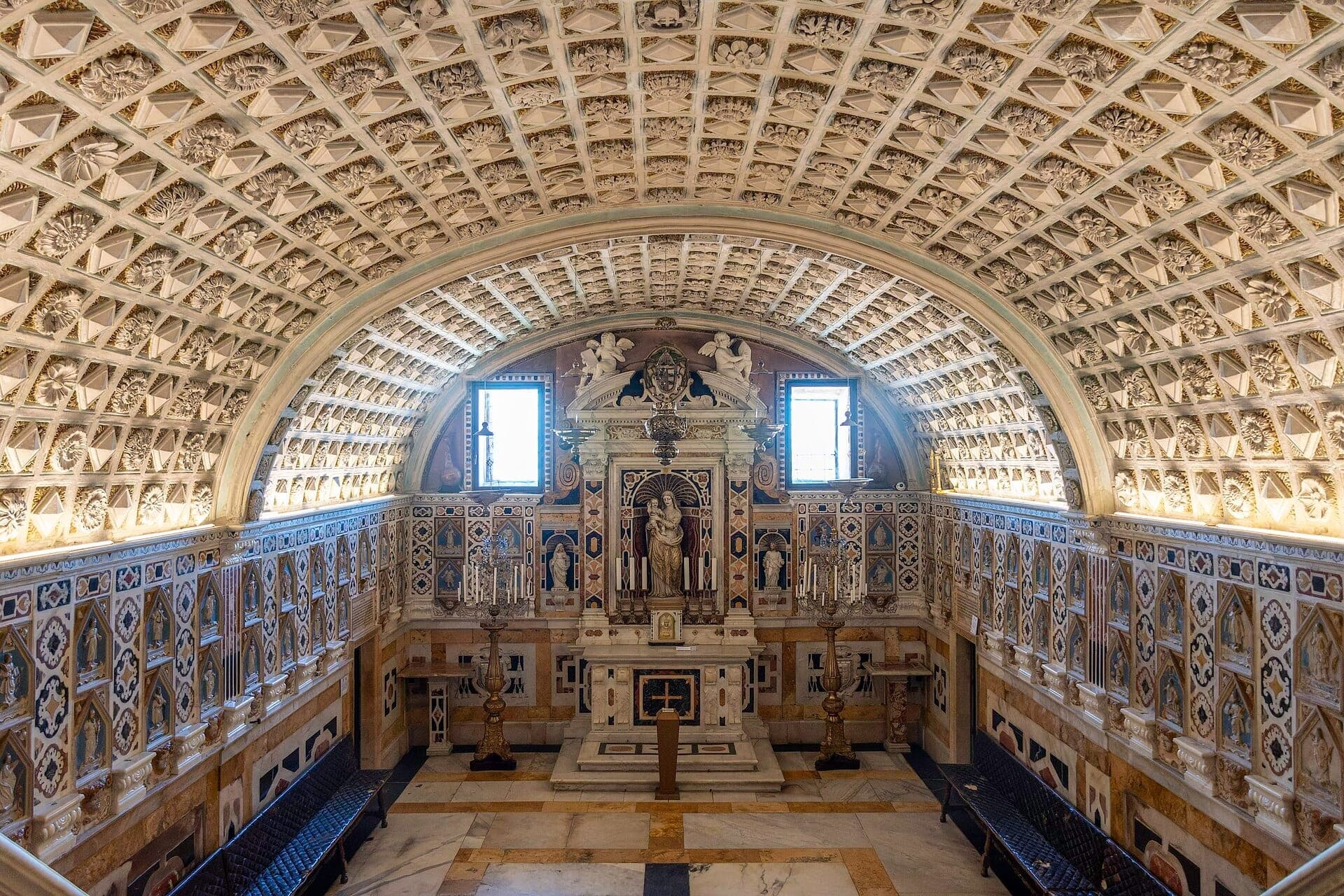 Techo abovedado intrincadamente decorado y altar de mármol en el interior de la Cattedrale di Santa Maria, Cagliari, con bancos simétricos y luz natural del día.