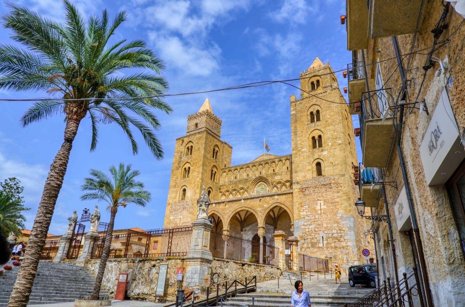 Fasad Katedral Cefalù di Sisilia dengan dua menara Norman, pohon palem, dan langit biru cerah yang dramatis, dilihat dari tangga masuk.