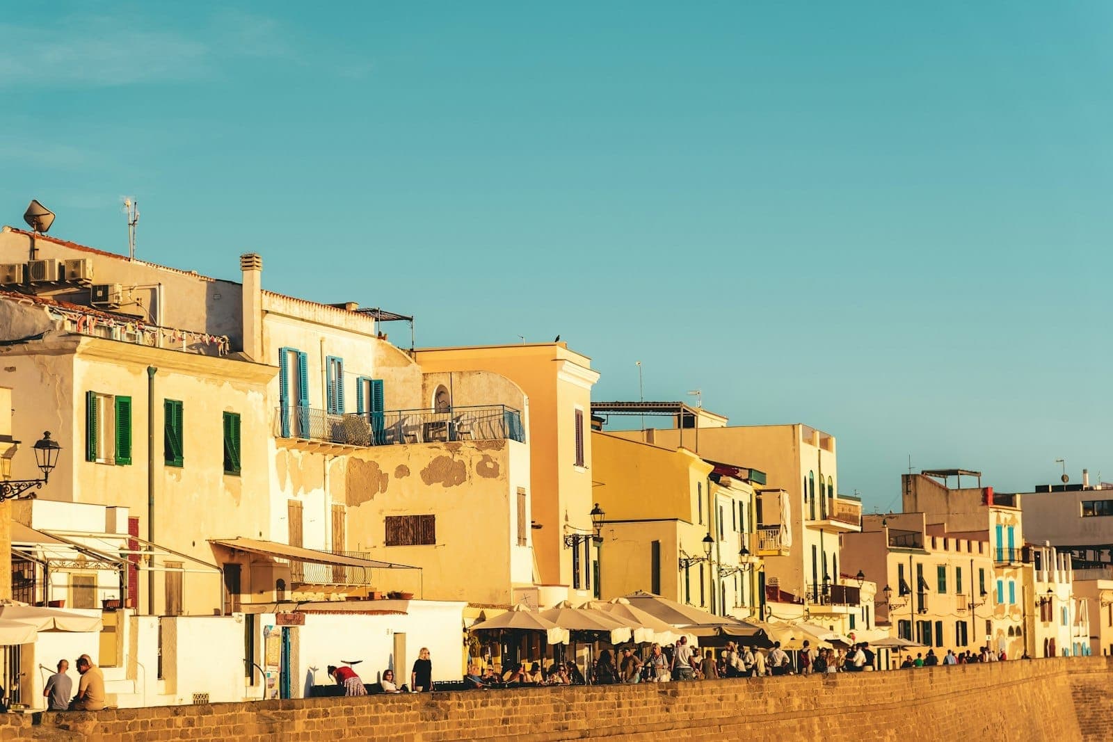 Edificios de color miel bañados por el sol con contraventanas azules bordean las murallas del centro histórico de Alghero bajo un cielo despejado, con gente reunida abajo.