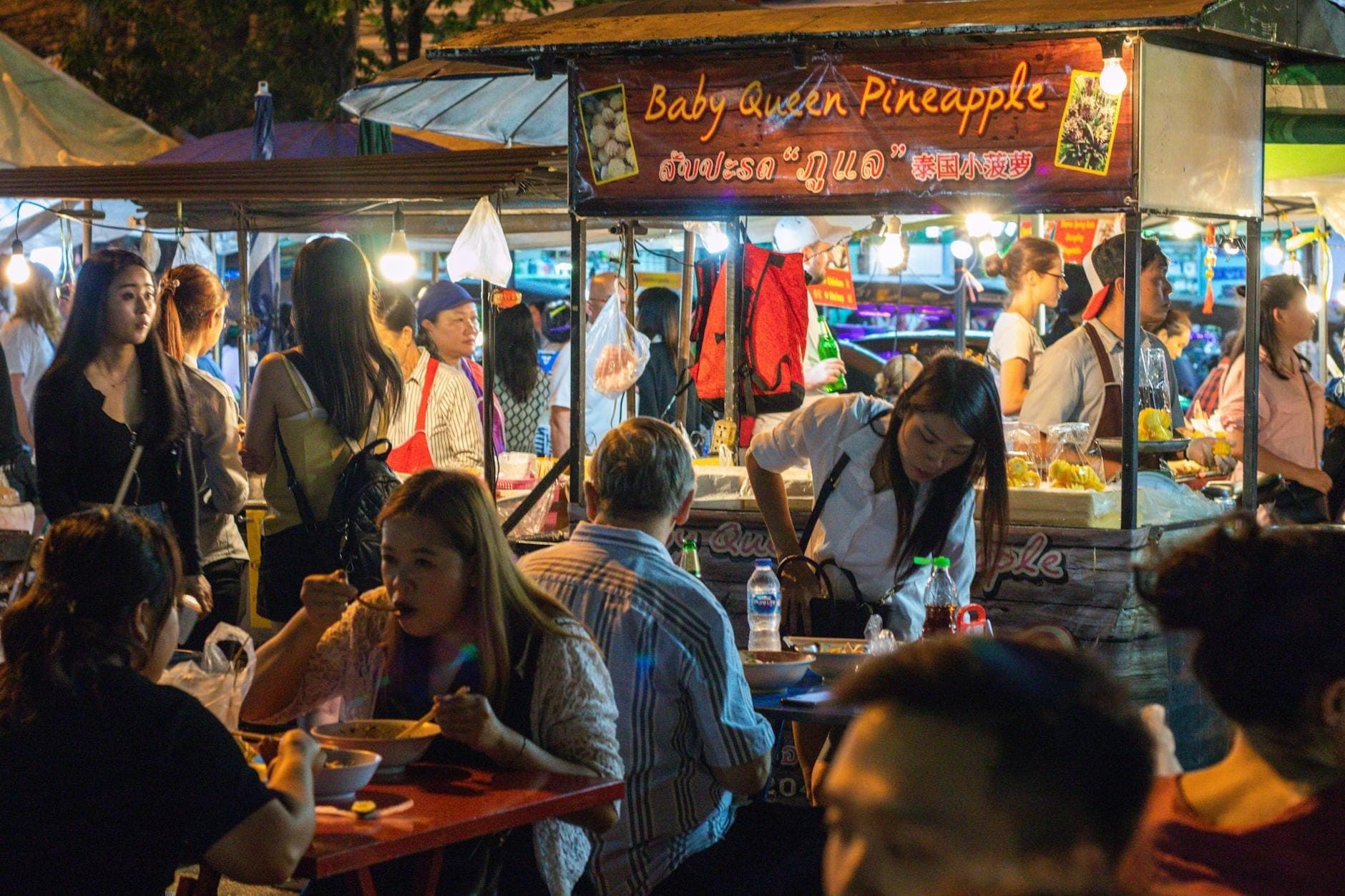 Belebter Nachtmarkt in Chiang Mai mit Menschenmassen, die Streetfood essen, und Verkäufern, die Gerichte unter hellen Marktlichtern servieren.