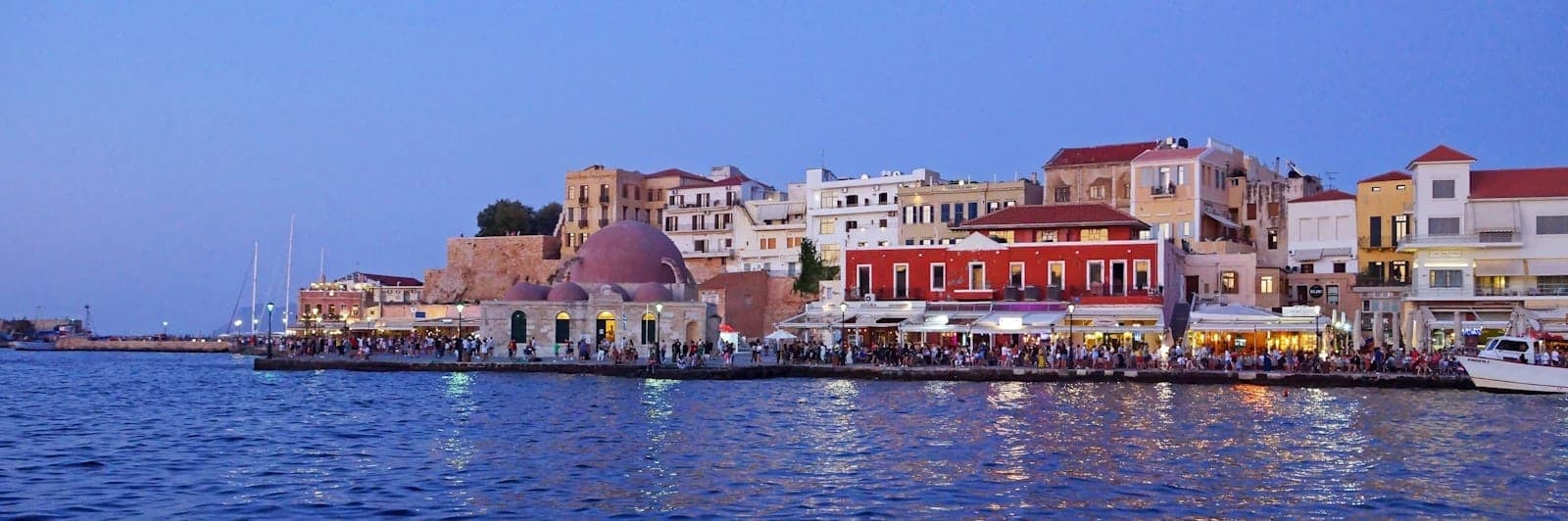 Abendliche Ansicht des Hafens von Chania mit bunten historischen Gebäuden, Restaurants und der bekuppelten Moschee, die sich im ruhigen Wasser unter einem violetten Dämmerungshimmel spiegelt.