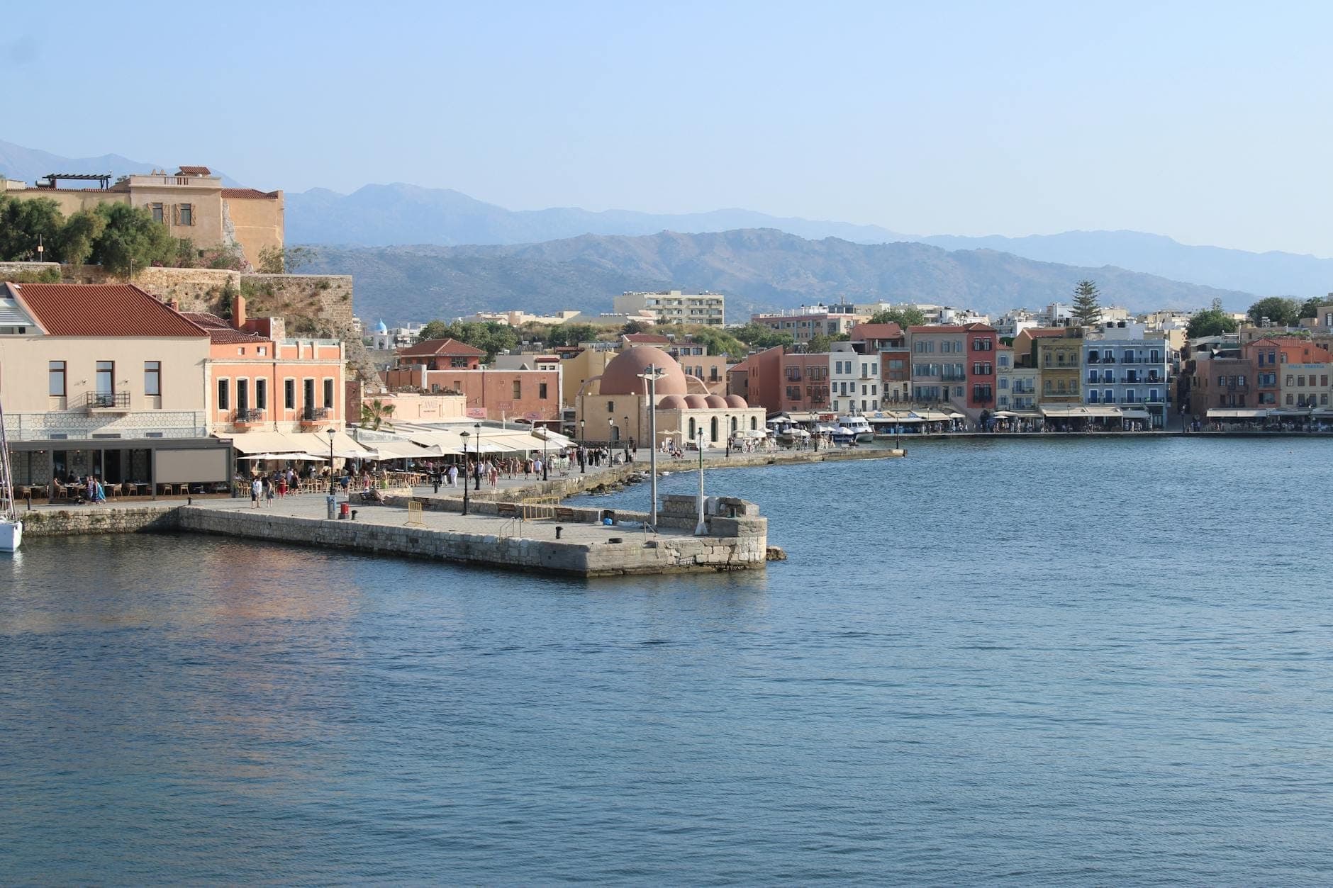 Bunte Hafenhäuser und die Kuppelmoschee im alten Hafen von Chania – blaues Meer, Bergkulisse und eine lebhafte, einladende Atmosphäre.
