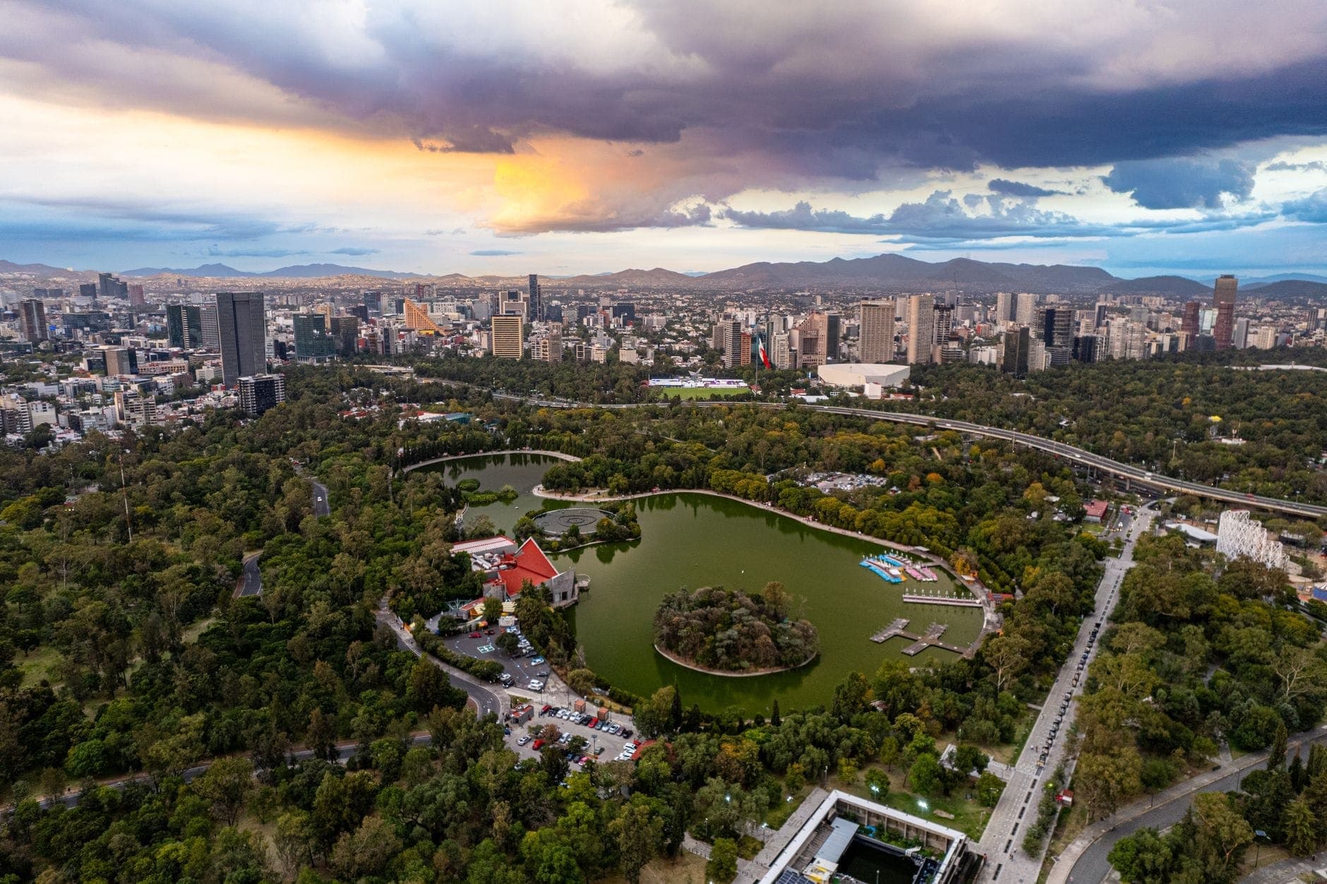 มุมมองทางอากาศของ Bosque de Chapultepec พร้อมต้นไม้เขียวชอุ่ม ทะเลสาบกลาง เรือสีสันสดใส และเส้นขอบฟ้าของ Mexico City ใต้ท้องฟ้ายามเย็นที่งดงาม