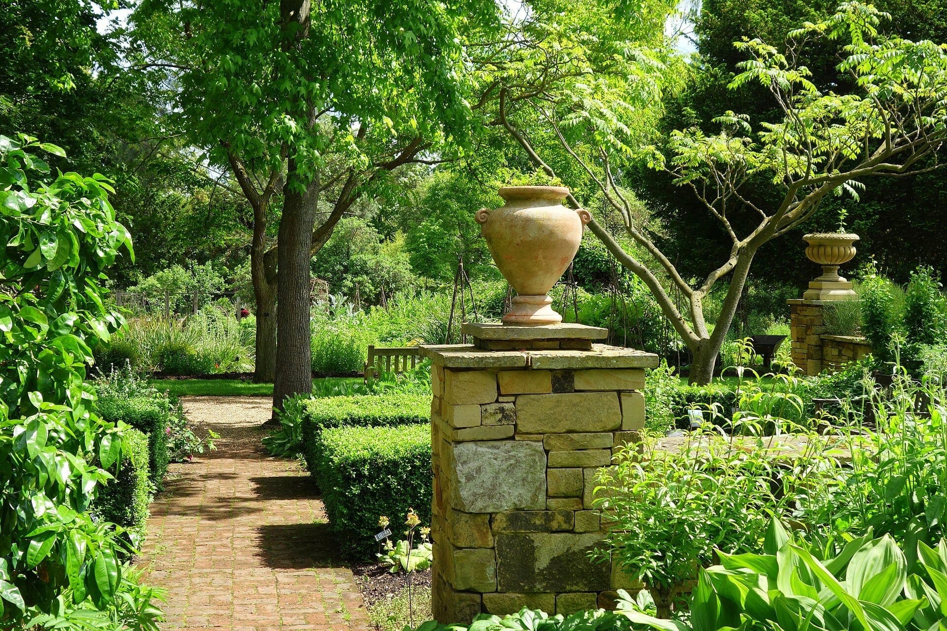 Słoneczny widok na Chelsea Physic Garden z kamiennymi ścieżkami, liściastymi drzewami, bujną zielenią i klasycznymi urnami na kamiennych postumentach.