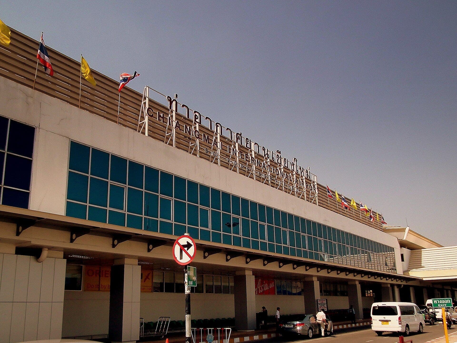 Außenansicht des Terminals des Chiang Mai International Airport mit Glasfenstern, Fahnenaufstellungen und geparkten Fahrzeugen im Abholbereich bei klarem Himmel.